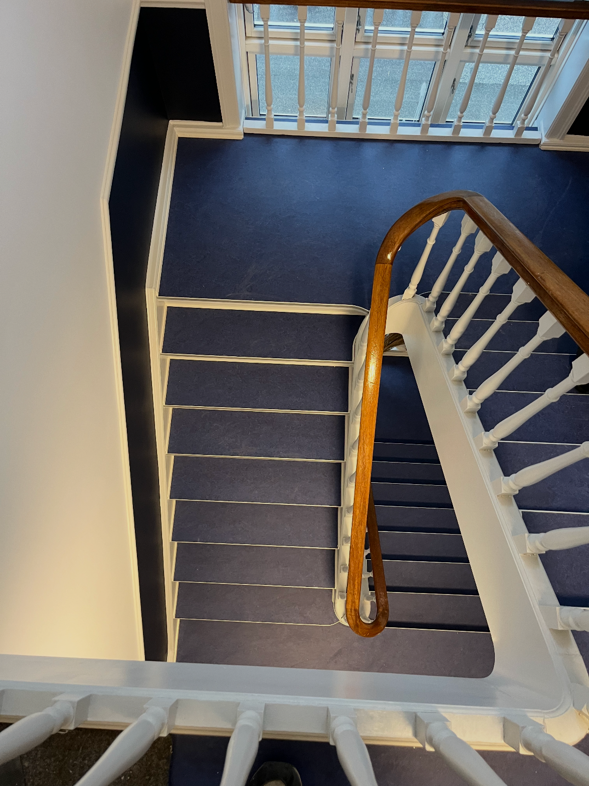 Top-down view of a staircase with navy blue carpet, white railing, and wooden handrail, leading to a landing with navy blue carpet and a glass door overlooking a patio.
