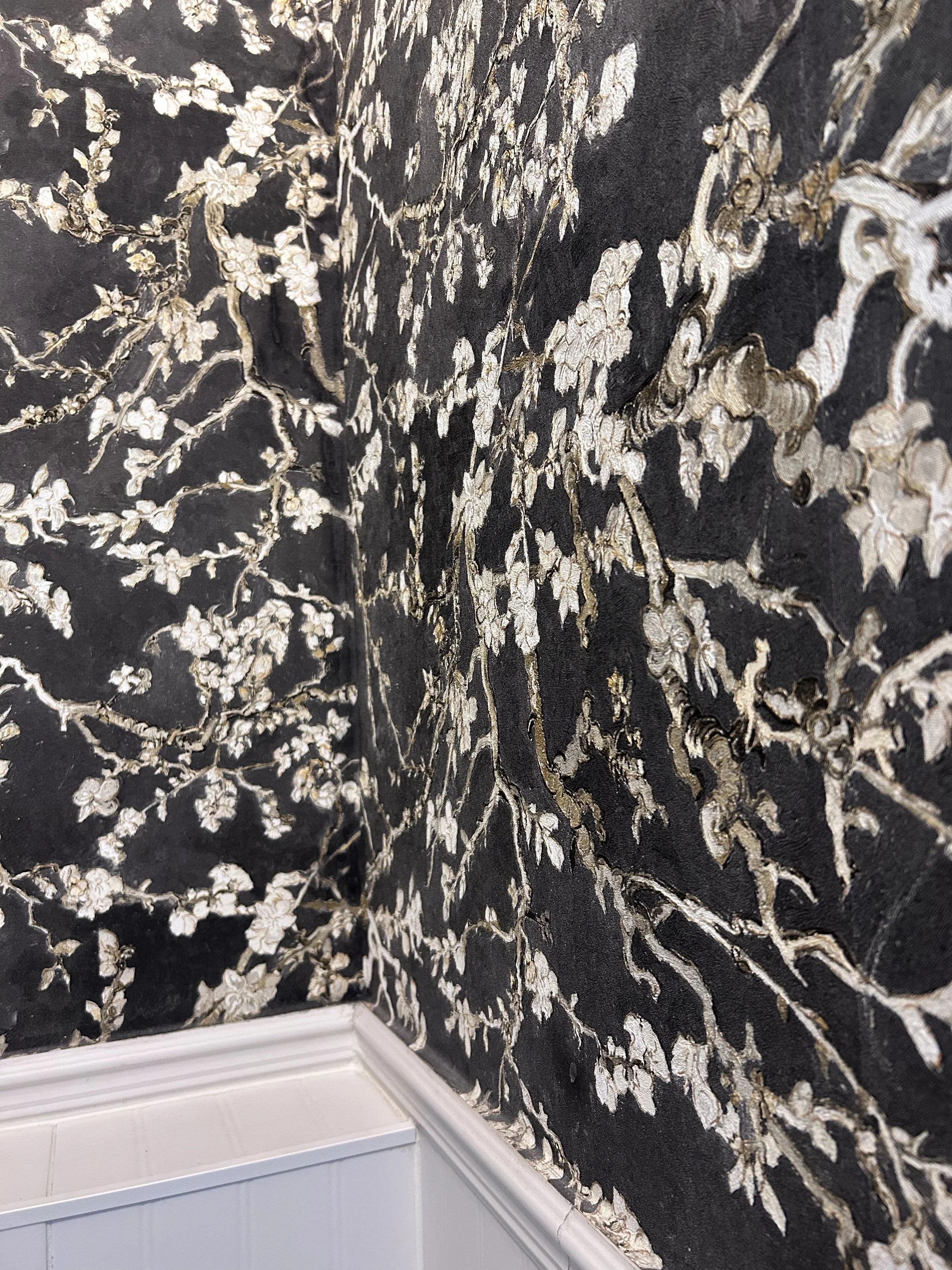 Close-up of floral wallpaper with black background and beige floral pattern, next to white wooden paneling and white baseboard at the bottom.