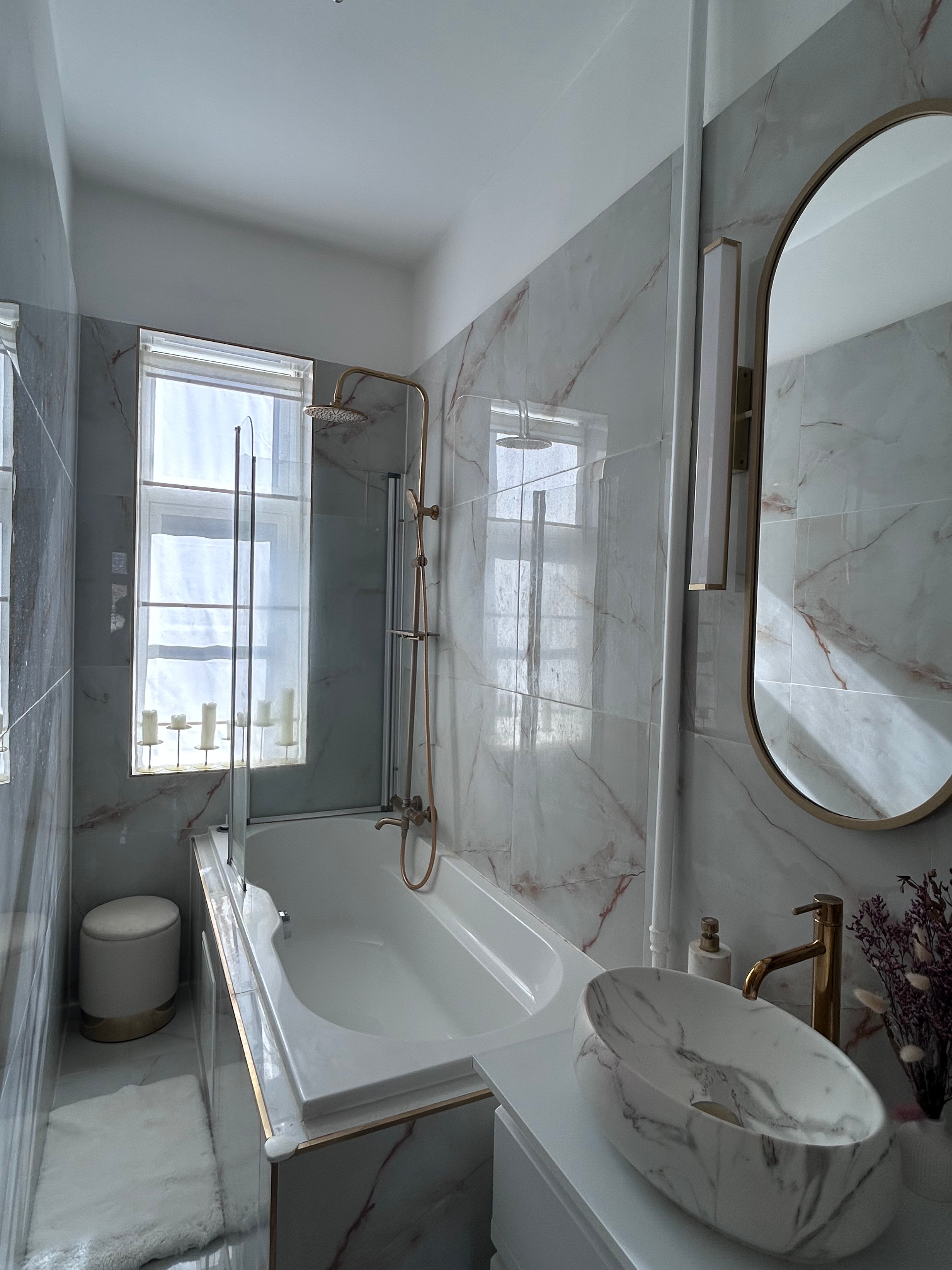 Modern bathroom with marble walls, a bathtub with a showerhead, a small round stool, a white rug, and a marble vessel sink with gold fixtures, a mirror, and a window with candles.