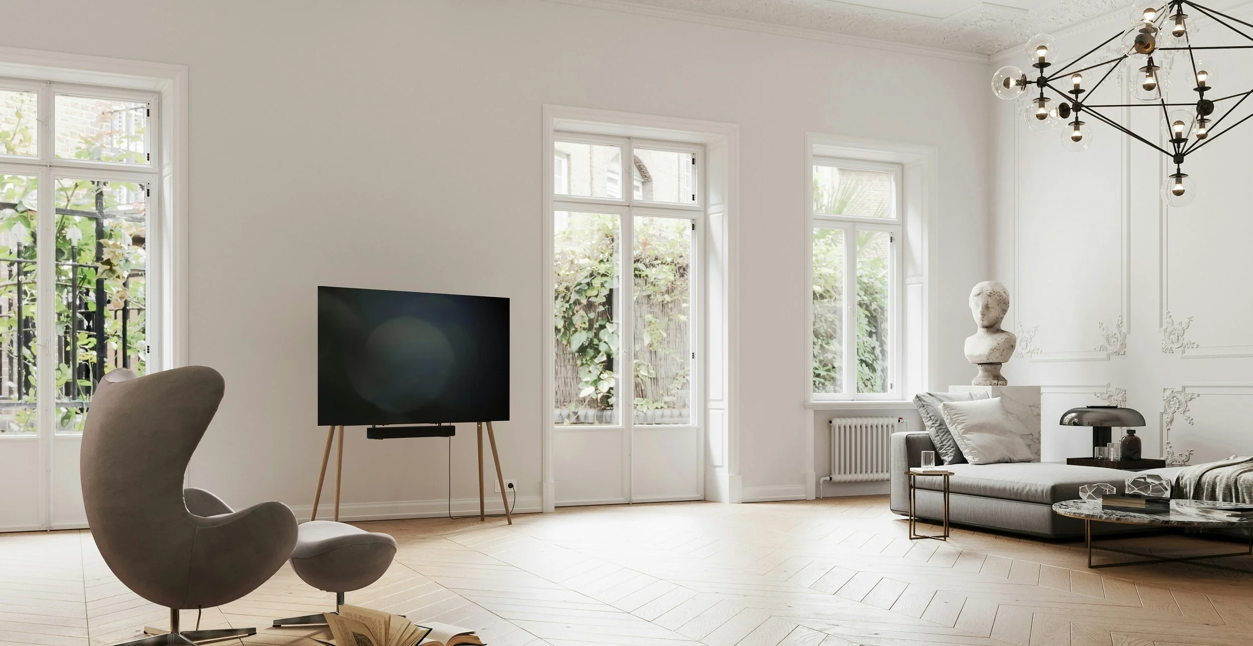Bright living room with white walls, large windows, a black TV on wooden stand, a modern gray armchair, a white sofa with pillows, a bust sculpture, a glass-topped coffee table, and a black chandelier.