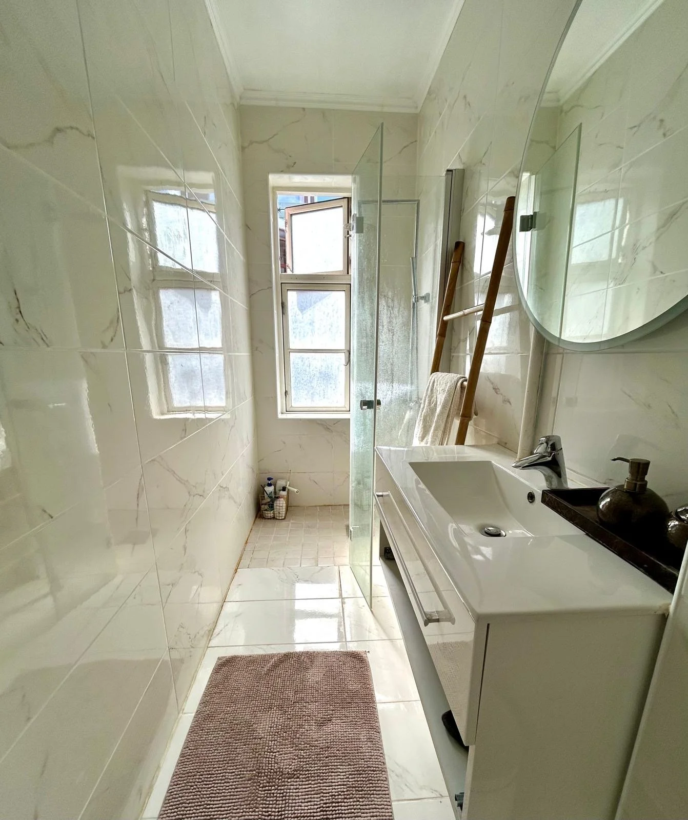 A small bathroom with cream-colored marble walls and floors, a window at the end, a white sink with a cabinet below, a mirror above, a black tray with soap and lotion, a towel hanging on a ladder-style rack, and cleaning supplies in a basket on the floor near the window.