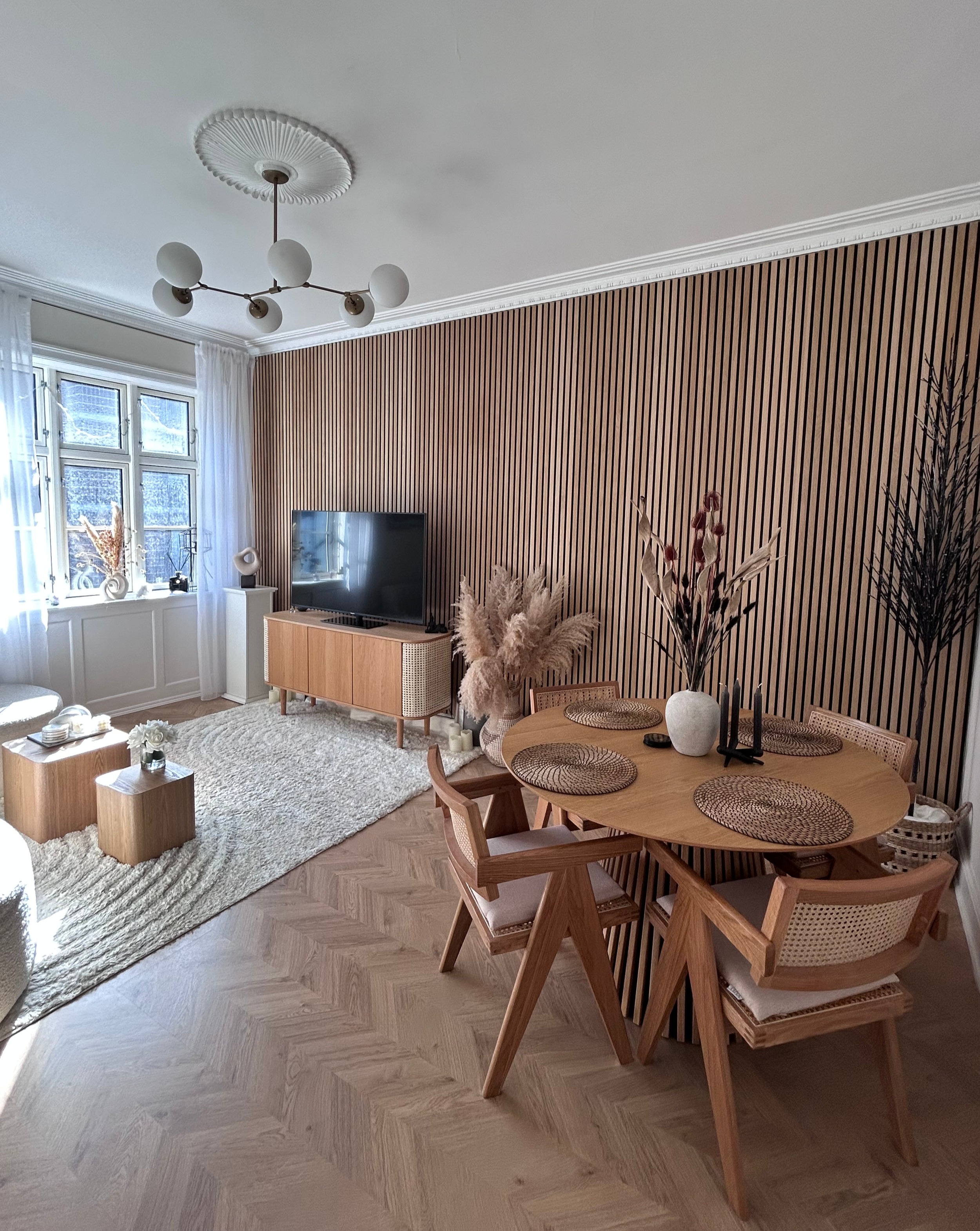 Living room with a wooden-paneled accent wall, a round dining table with six wicker placemats and vases, a TV on a wooden stand, large window with white curtains, and a beige rug.