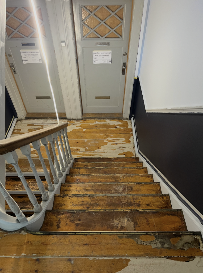 View of a staircase with worn wooden steps leading to a door inside a house. The door has a small mail slot and a decorative upper panel. The wall beside the staircase is painted black, and the area near the door shows signs of wear and peeling paint.