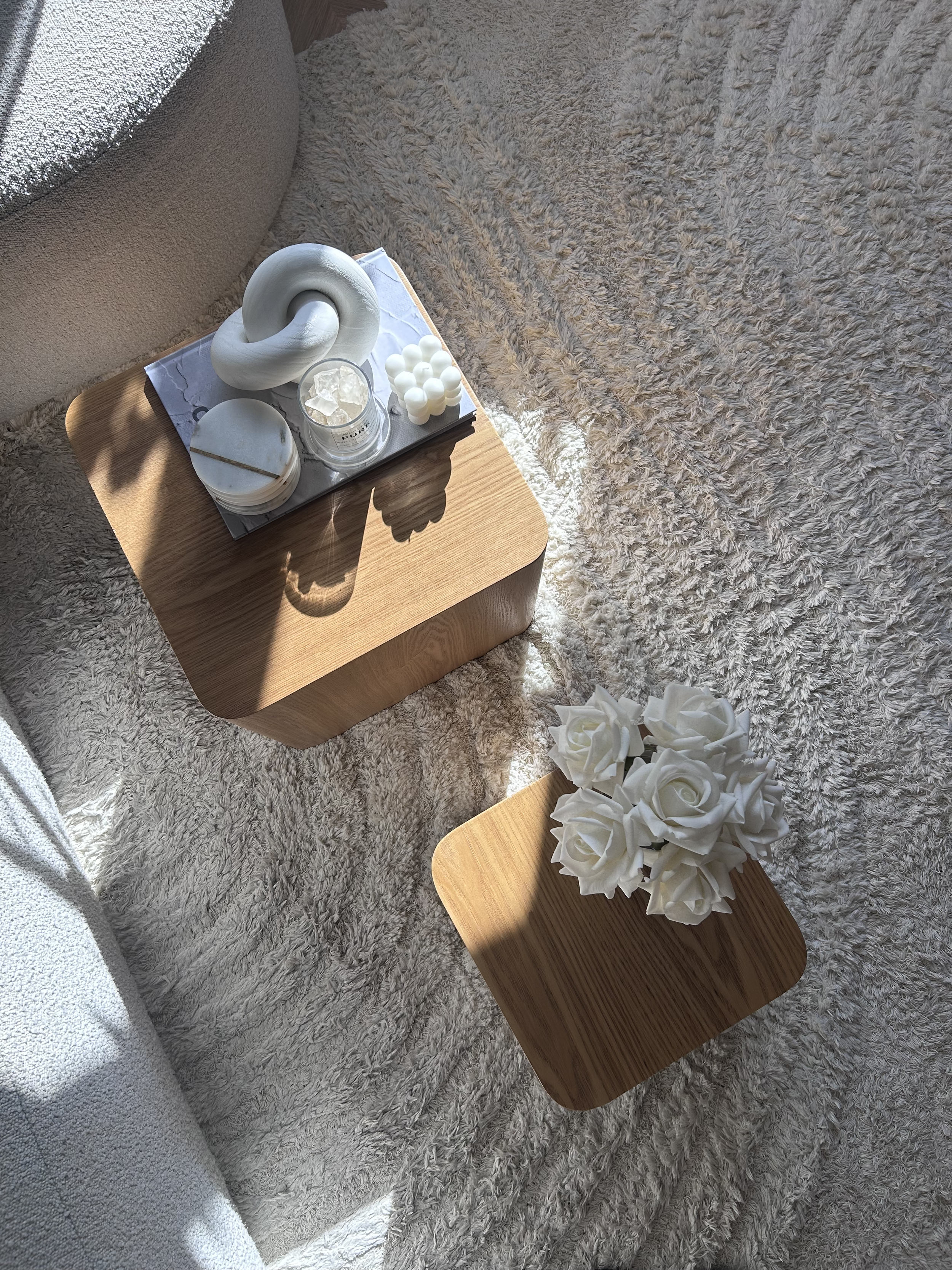 Two wooden tables with decorative items: one with white candles, a white sculpture, and a candle holder, the other with a bouquet of white roses, set on a textured beige carpet with sunlight streaming in.