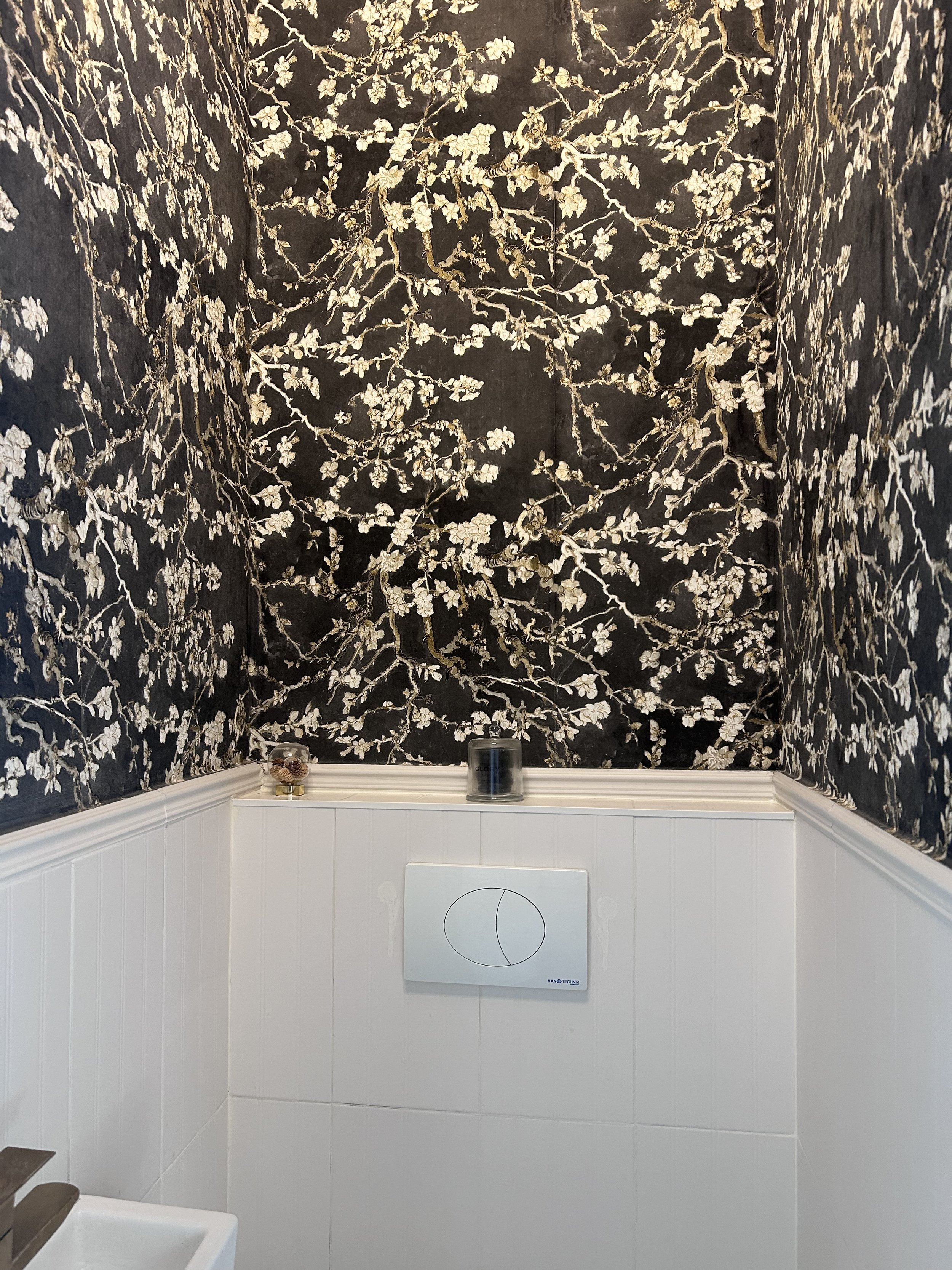 A wall in a bathroom with black and white floral wallpaper on the upper walls and white paneling on the lower part, with a dual-flush toilet button and small decorative items on the ledge.