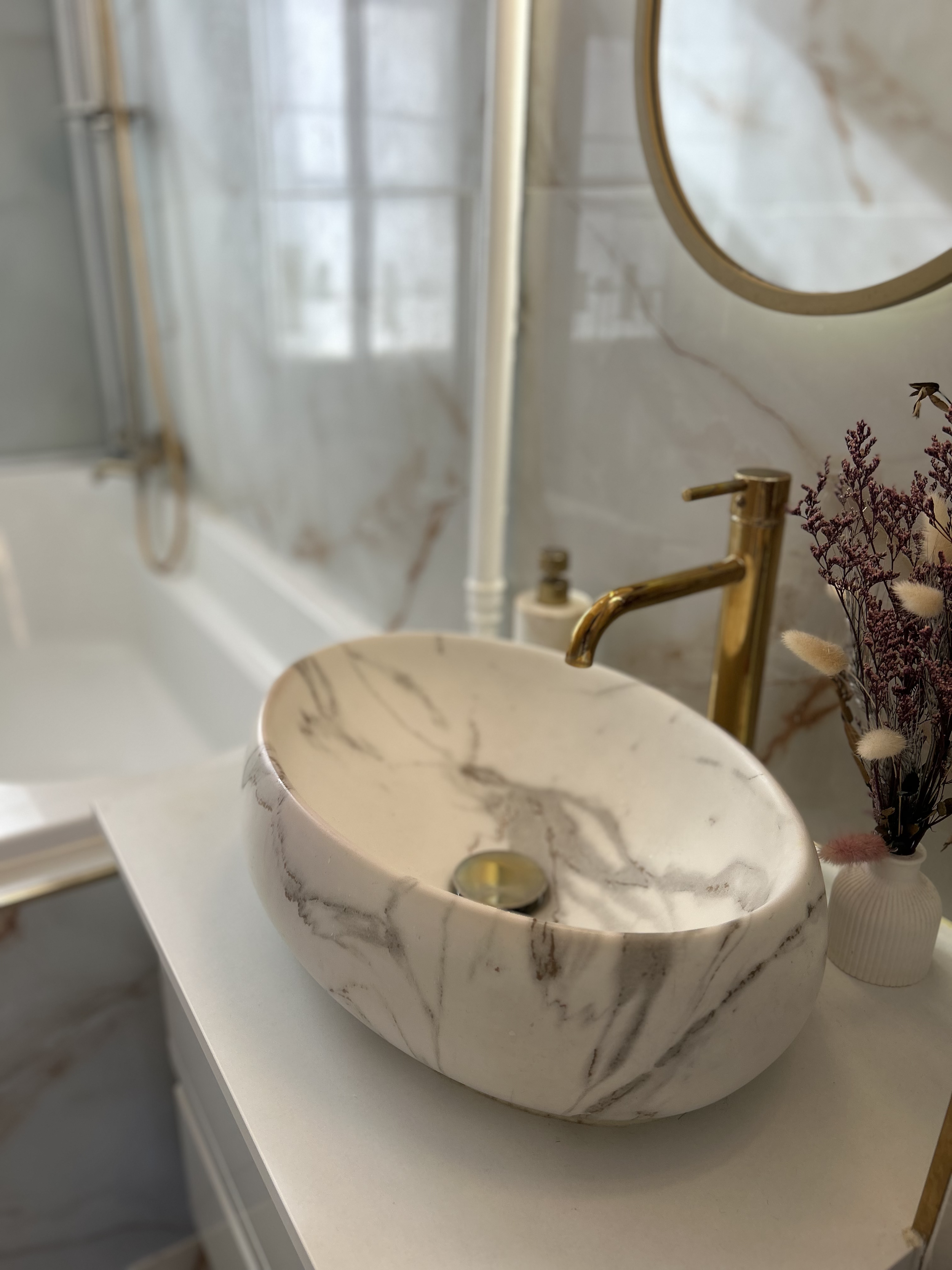 Marble bathroom sink with gold faucet next to a vase of dried flowers in a modern bathroom.
