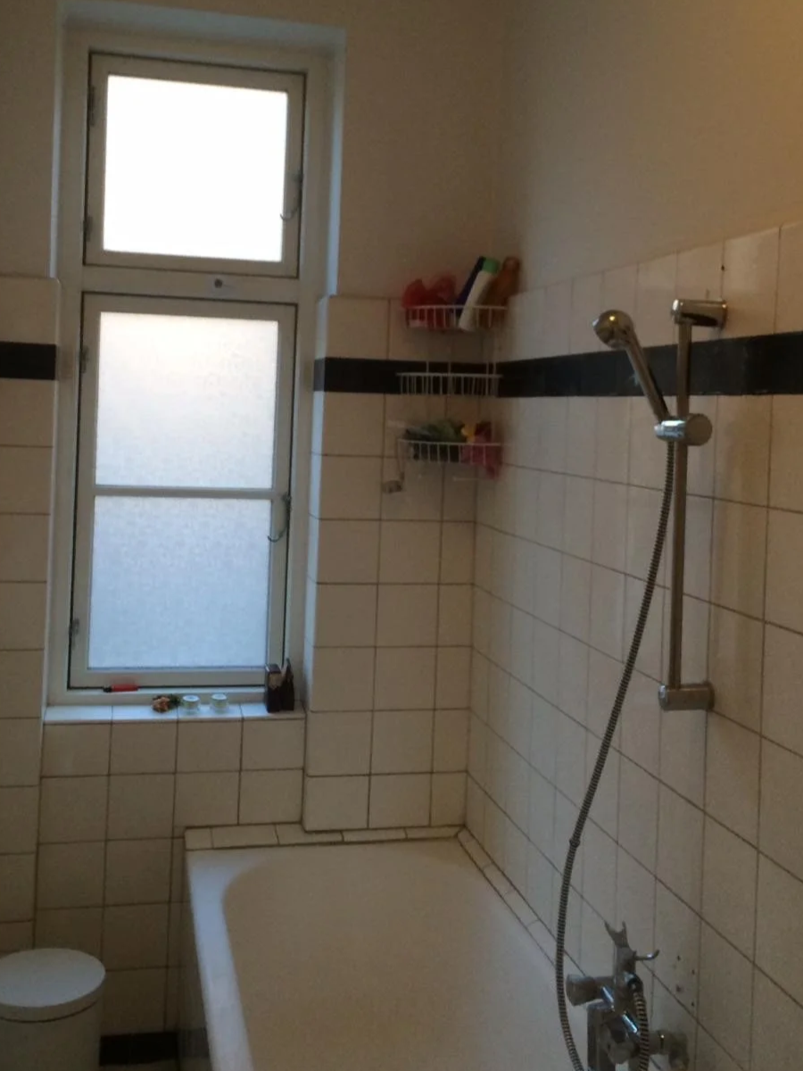 A bathroom with a frosted window, tiled walls with a black stripe, a bathtub, a showerhead with an adjustable hose, and small wire shelves holding toiletries.