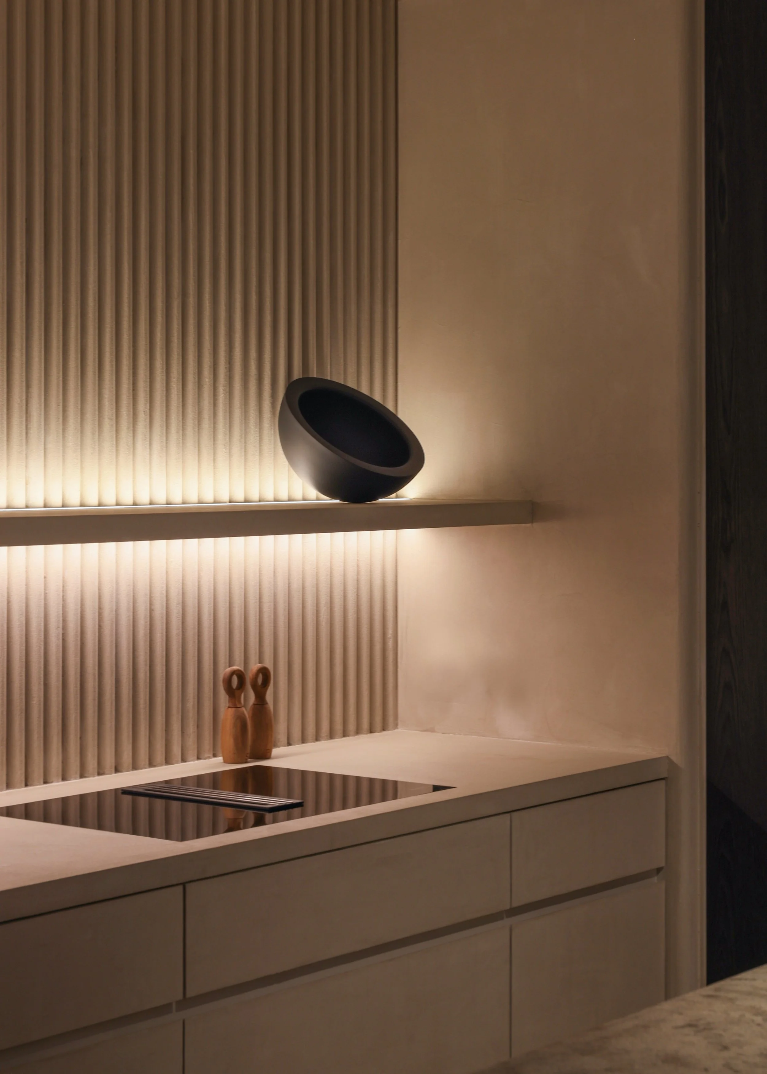 Modern kitchen with beige cabinetry, a black bowl on a shelf, and two wooden decorative objects on the counter, with warm lighting.