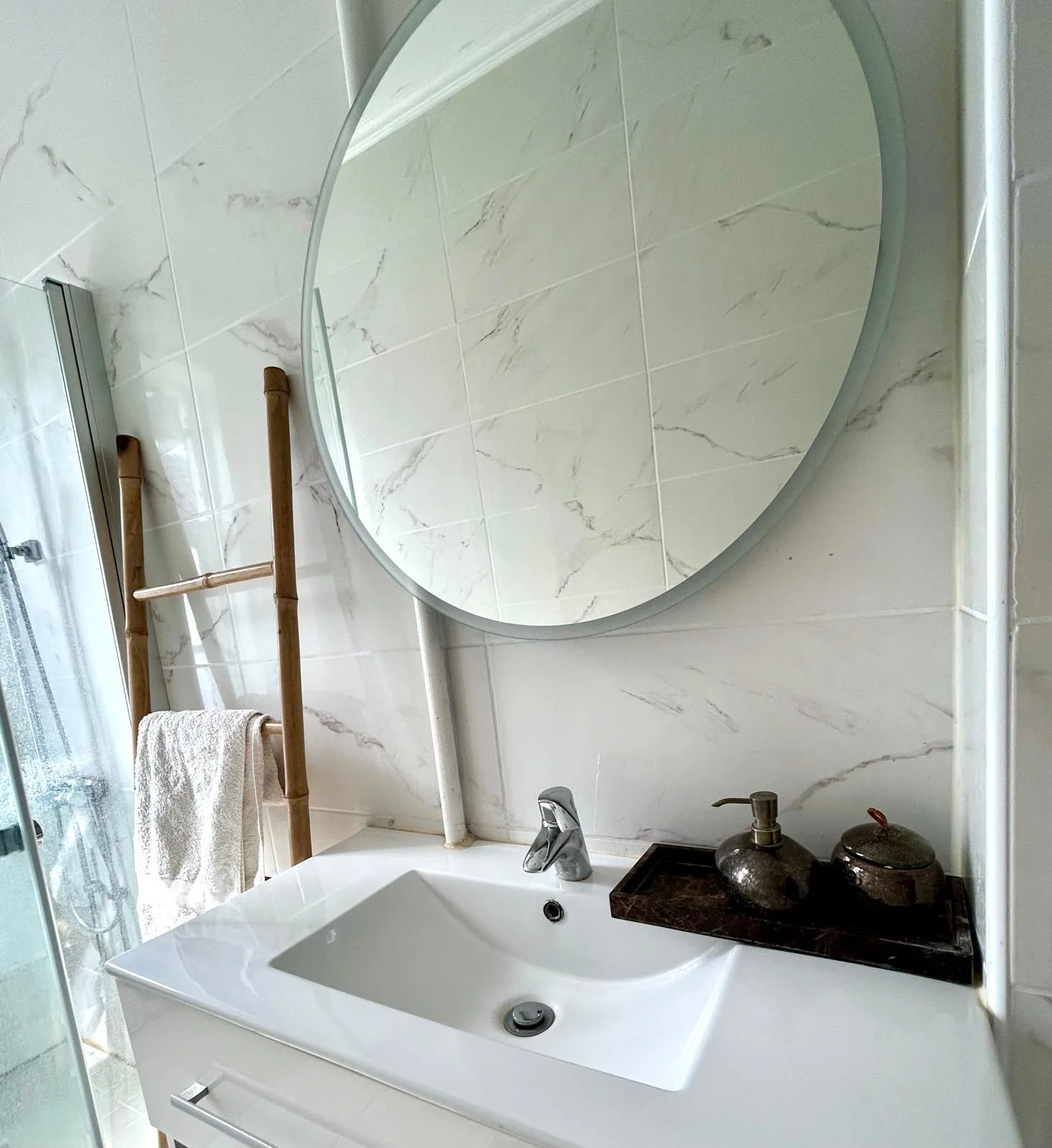 A modern bathroom with white marble walls, a round mirror above a white sink, and a bamboo towel ladder with a white towel. There is a black tray with two decorative containers on the sink.