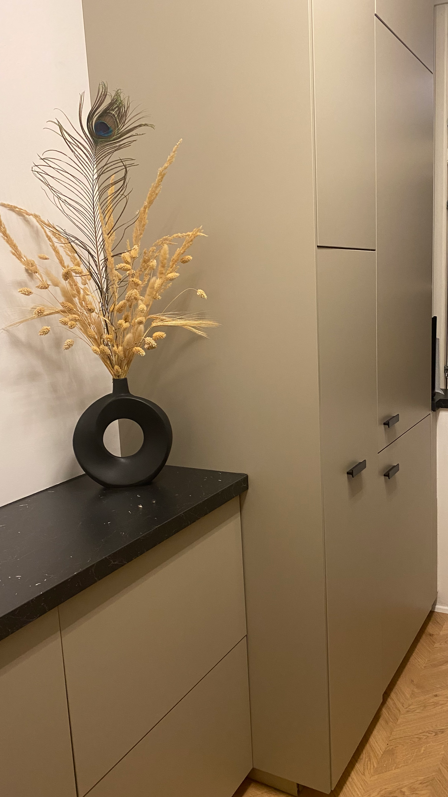 A modern interior space with beige cabinetry, a black countertop, and a decorative black vase with peacock feather and dried plants.