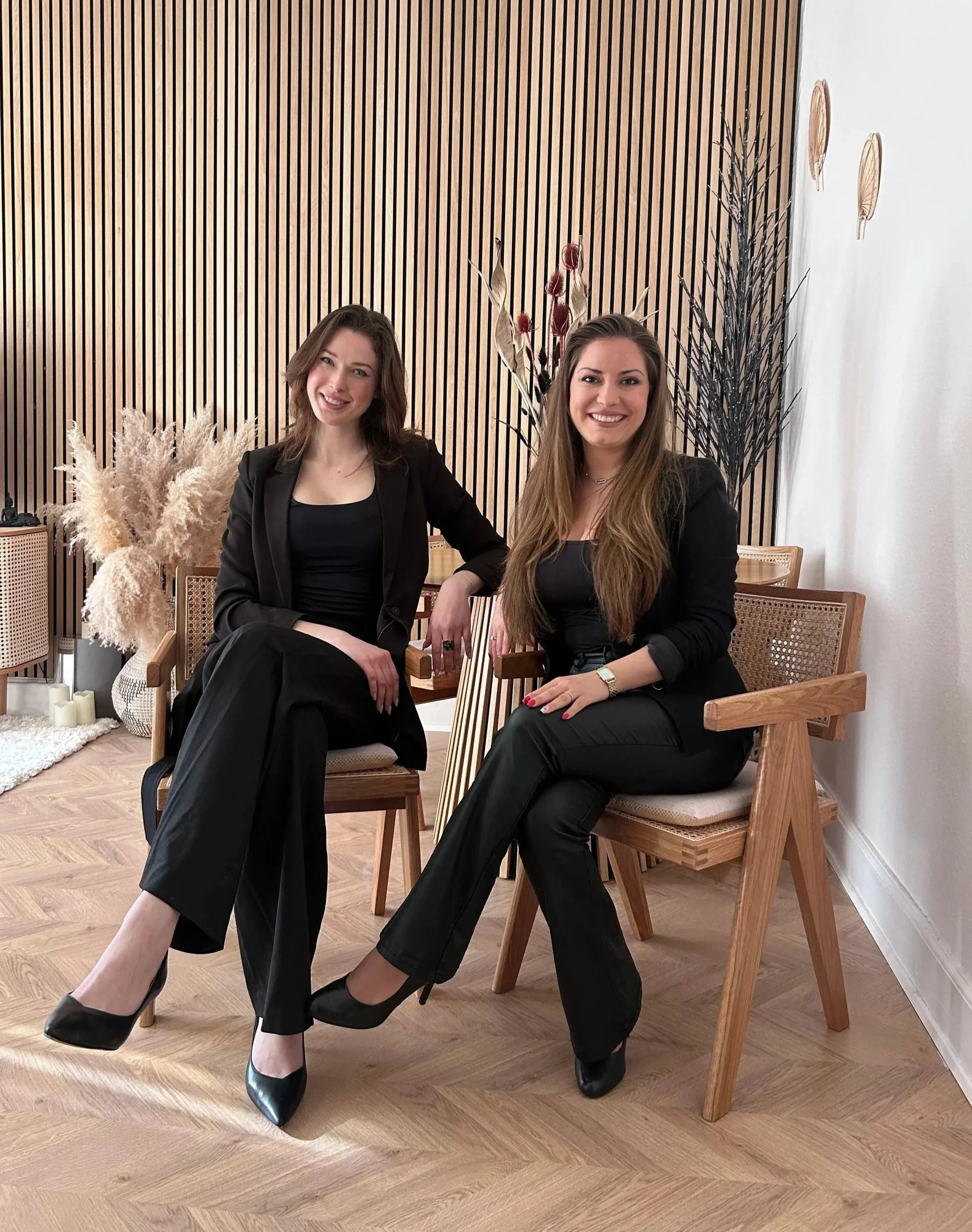 Two women in black outfits sitting on wooden chairs, smiling at the camera, in a room with wooden decor and dried plants in the background.