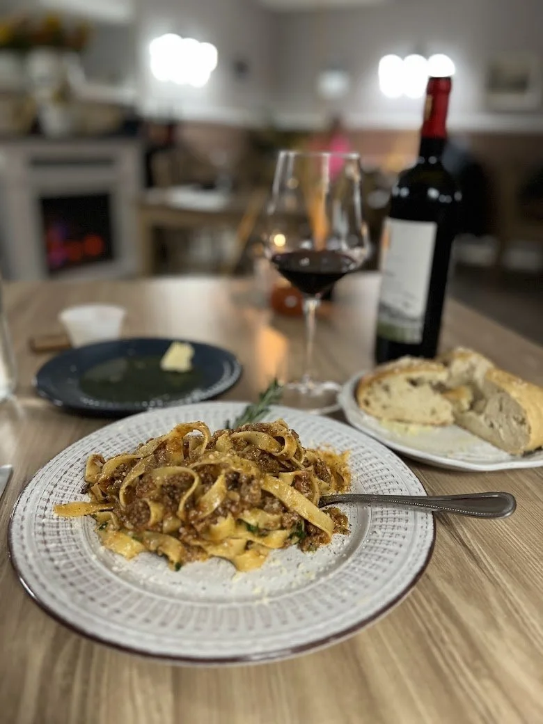 A dinner table with a plate of pasta, a glass of red wine, a bottle of wine, slices of bread, a small dish, and a green plate with a piece of cheese, in a cozy kitchen setting.