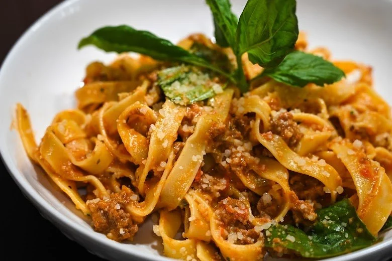 A bowl of cooked fettuccine pasta with meat sauce, garnished with fresh basil leaves and grated cheese.