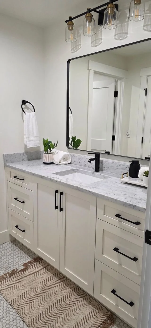Bathroom vanity with white cabinets, marble countertop, black faucet, large mirror, black light fixture, white towels, potted plant, soap dispenser, and a gray and beige striped rug.