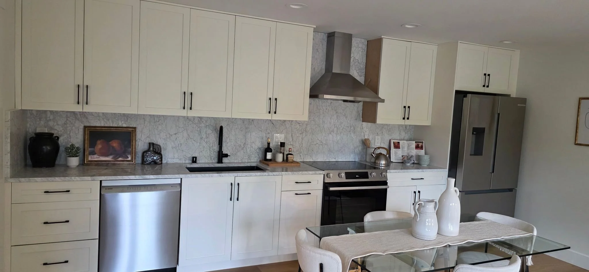 Modern kitchen with white cabinets, marble backsplash, stainless steel refrigerator, oven, and dishwasher, with a glass dining table and white chairs.