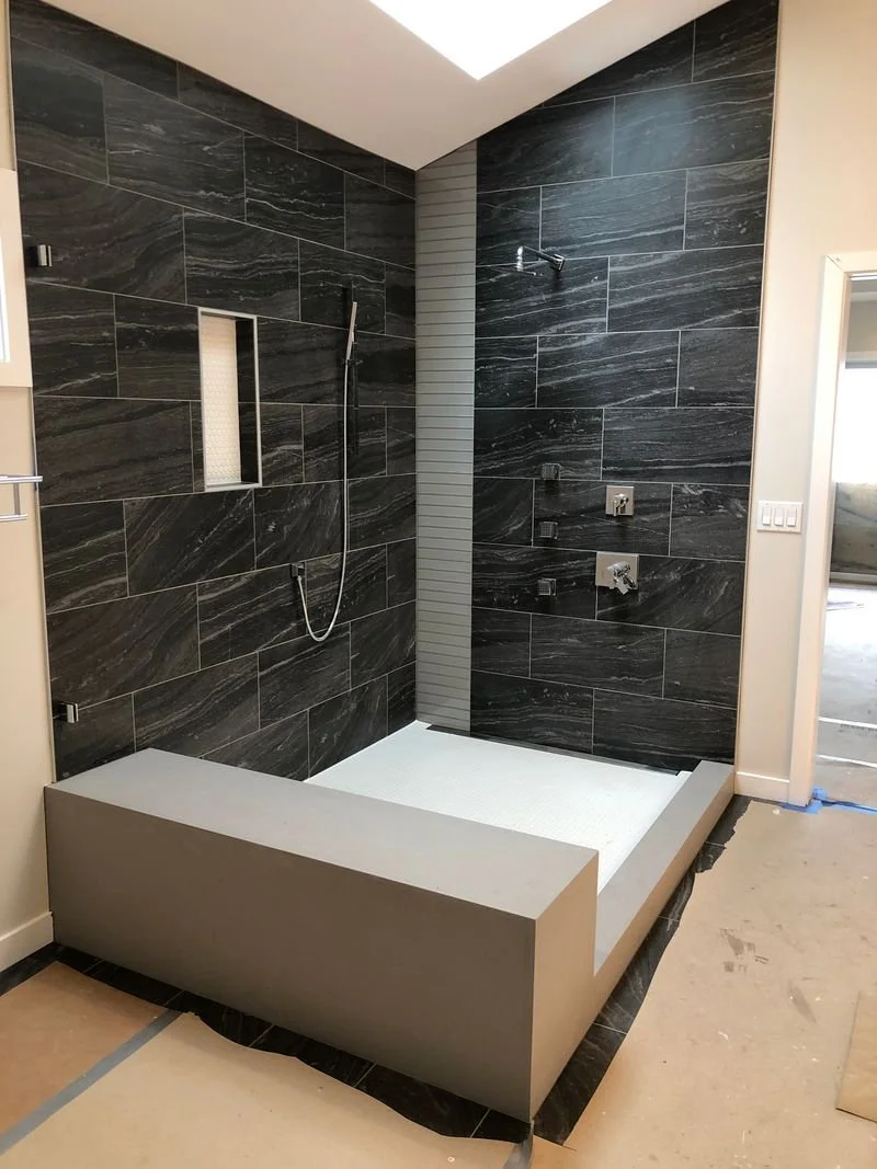 Modern walk-in shower with black marble tiles, a built-in niche, a rain shower head, a handheld shower, and multiple shower controls, with a raised light gray platform and partial view of surrounding beige walls.