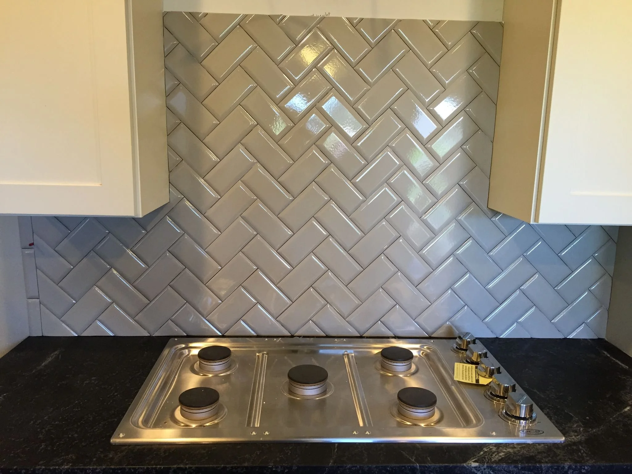Stainless steel stovetop with five burners and five control knobs, installed on a black granite countertop. The backsplash features light gray, glossy, arranged in a herringbone pattern tile.