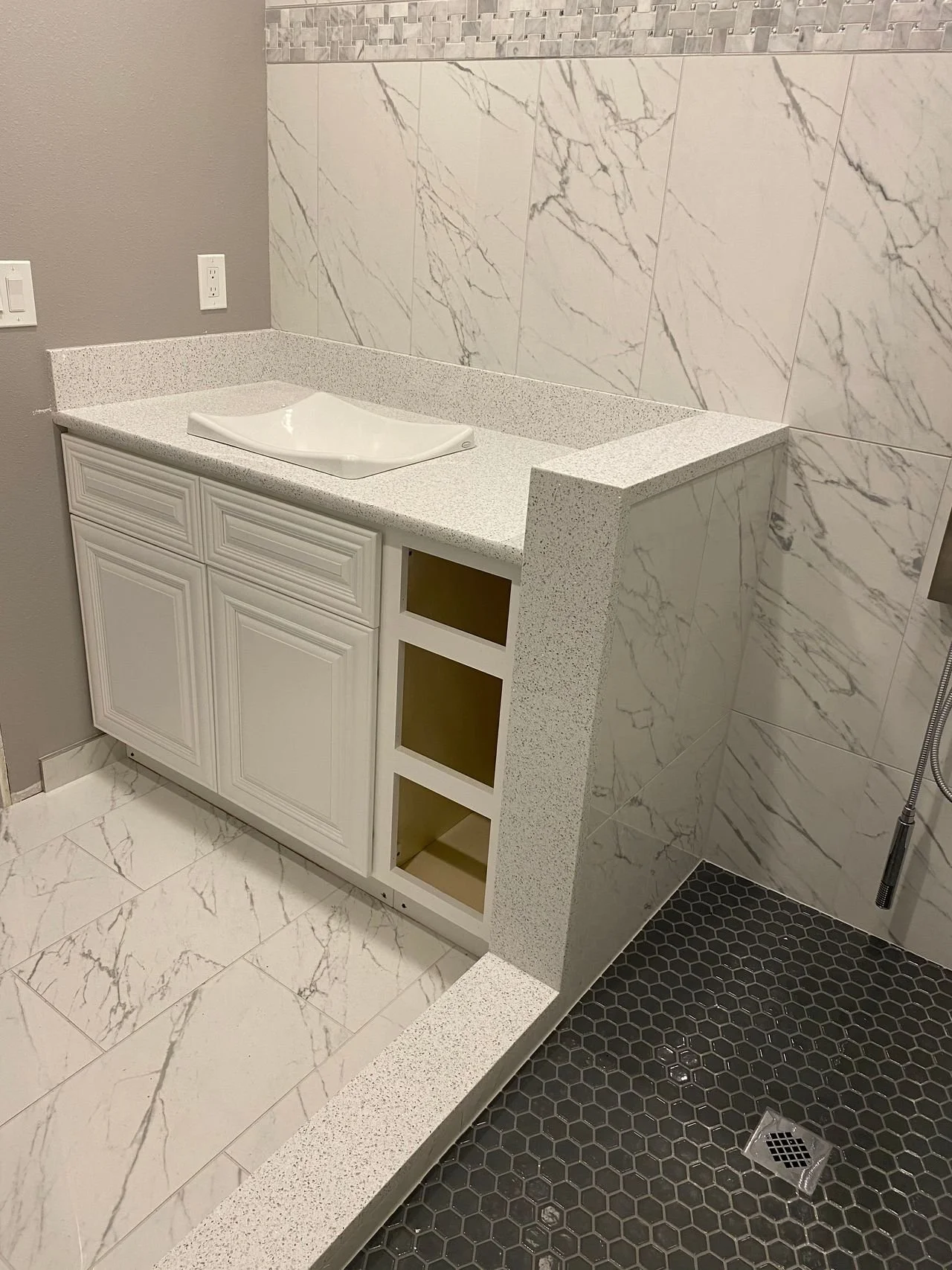 Bathroom vanity with a white cabinet and a speckled white countertop, located next to a tiled wall and a walk-in shower with black hexagonal tiles.