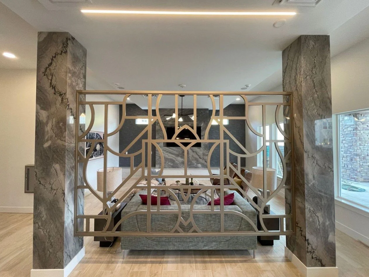 Modern open living room with a decorative metal room divider, a gray sofa with red pillows, marble pillars, large windows, and minimalist ceiling lighting.