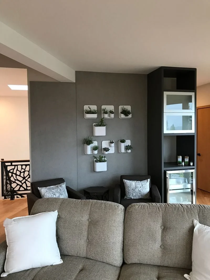 Living room corner with beige sofa, two gray armchairs, a small side table, and a wall with decorative white planters holding green plants. A tall black cabinet with glass doors is next to the wall, and a wooden door is visible on the right.