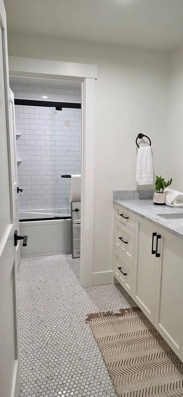 A modern bathroom with white subway tile shower, black hardware, white vanity with marble countertop, towel ring with towel, potted plant, and beige woven rug.