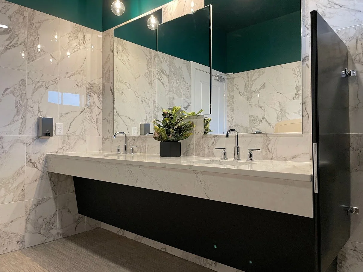 Modern bathroom with marble countertops and walls, featuring a large mirror, two sinks with chrome faucets, a black planter with green plants, and teal ceiling lighting.