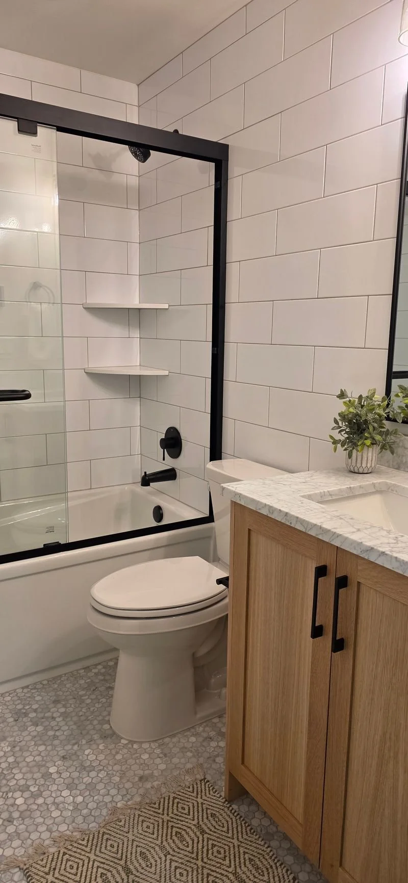A modern bathroom with white subway tile walls, a walk-in shower with black framing and fixtures, a white toilet, and a wooden vanity with a marble countertop and a potted plant.