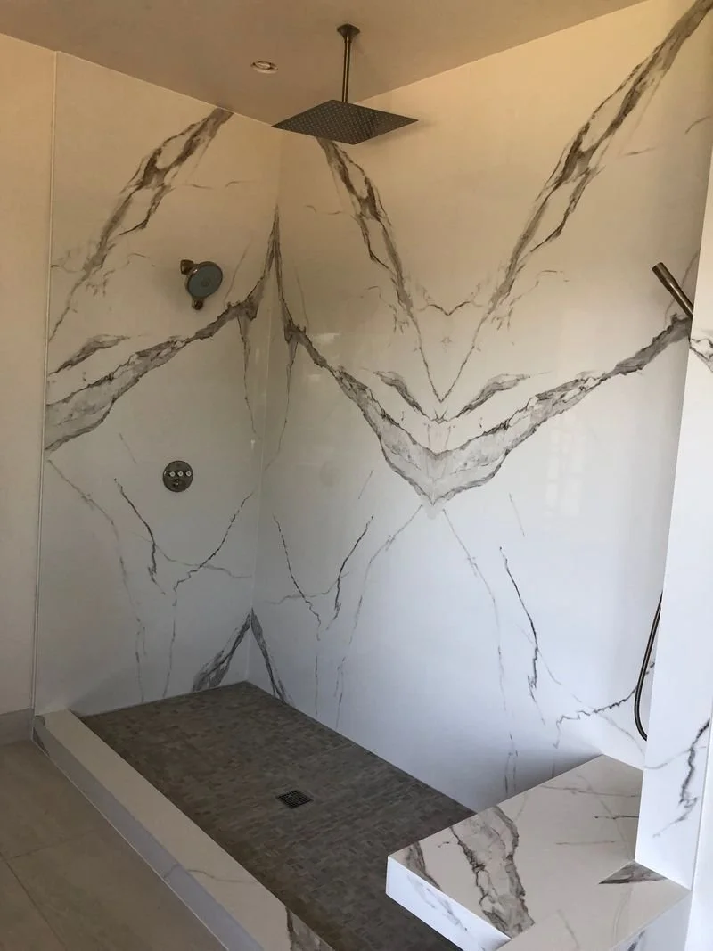 A modern walk-in shower with white marble walls featuring gray and black veining, a ceiling-mounted rain shower head, a small wall-mounted shower head, and a built-in marble bench.