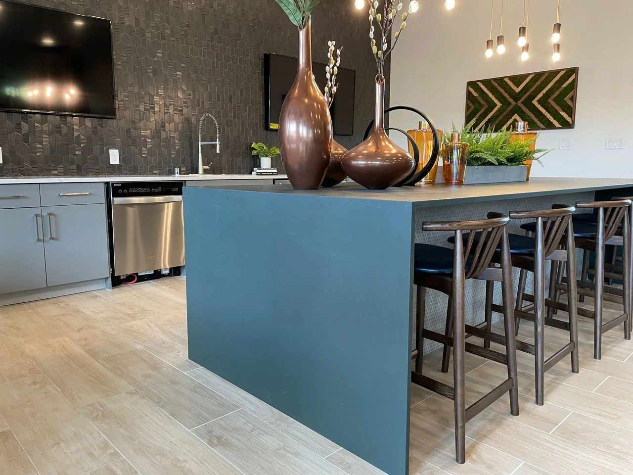 Modern kitchen with a blue island, four wooden barstools, black and metallic vases, green plants, a textured black backsplash, and pendant lighting