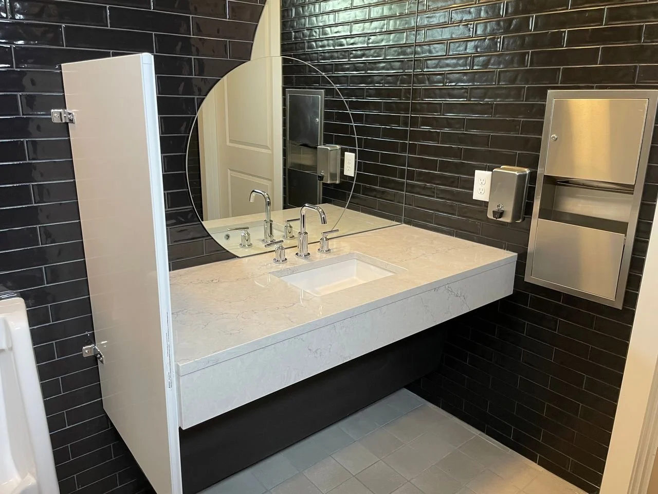 Bathroom sink with a round mirror, black tiled wall, white marble countertop, and a stainless steel paper towel dispenser.