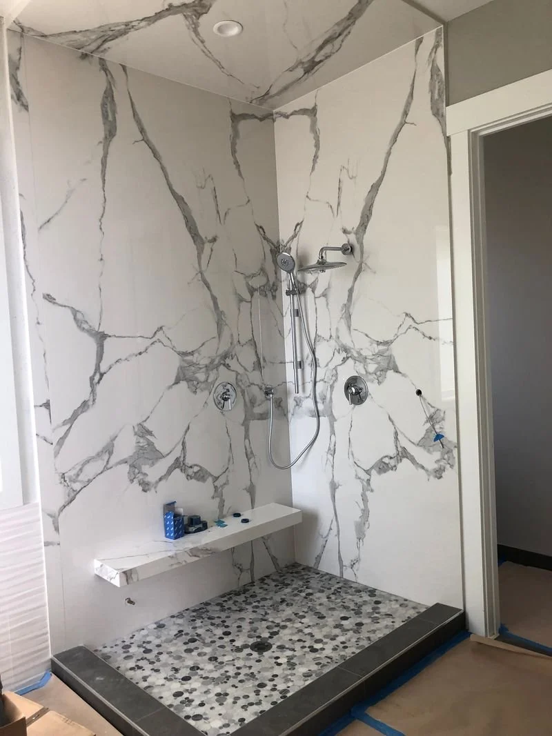 A modern walk-in shower with white marble walls featuring gray veining, pebble stone mosaic flooring, and a built-in bench. The shower has a handheld showerhead and two control knobs, with a corner shelf holding toiletries.