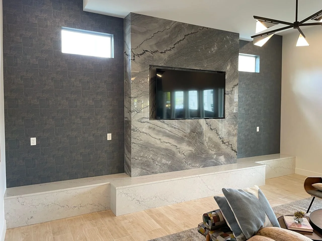 Modern living room with a wall-mounted flat-screen TV, gray patterned wallpaper, gray and beige marble accents, and a small window letting in natural light.