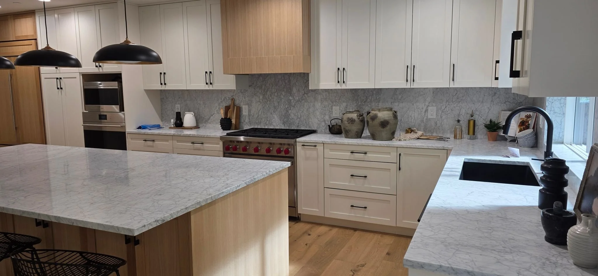 Modern kitchen with white and wood cabinets, marble countertops, a black sink, stainless steel oven, and decorative vases and plants by the window.