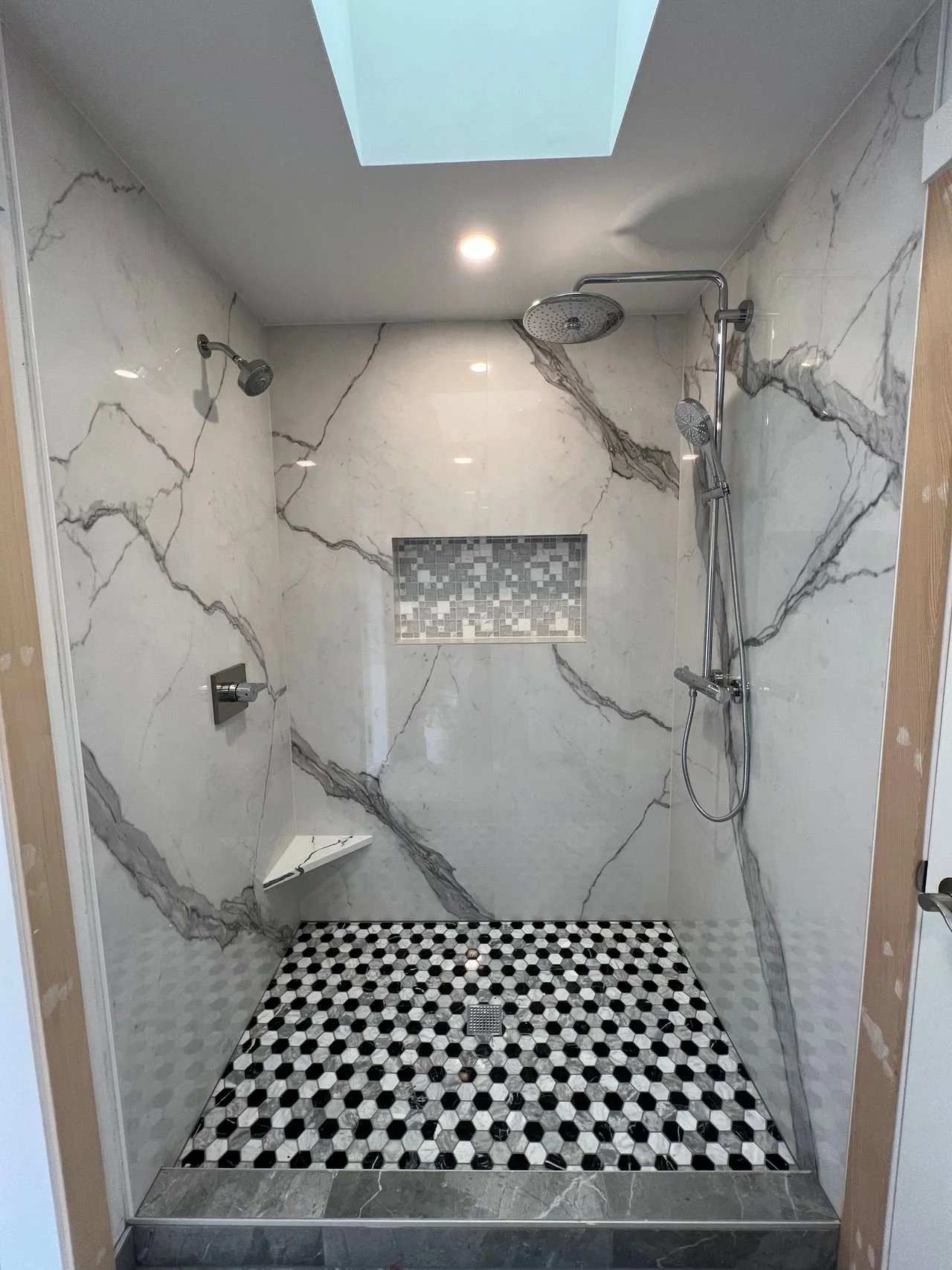 A spacious modern shower with white marble walls featuring dark gray veining, a built-in rectangular stone tile niche, and black, white, and gray patterned small hexagonal floor tiles. The shower has two showerheads, one fixed and one handheld, with 