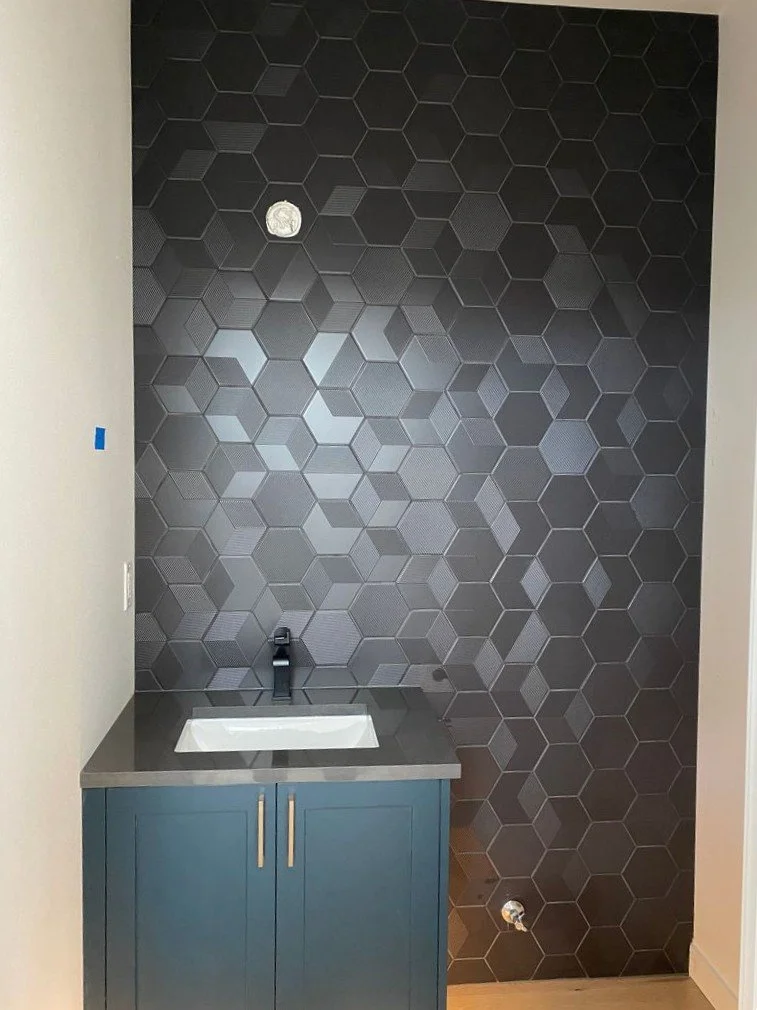 Small utility sink cabinet with a black faucet, mounted on a wall with a black hexagonal tile pattern, and an exposed water pipe connection below.