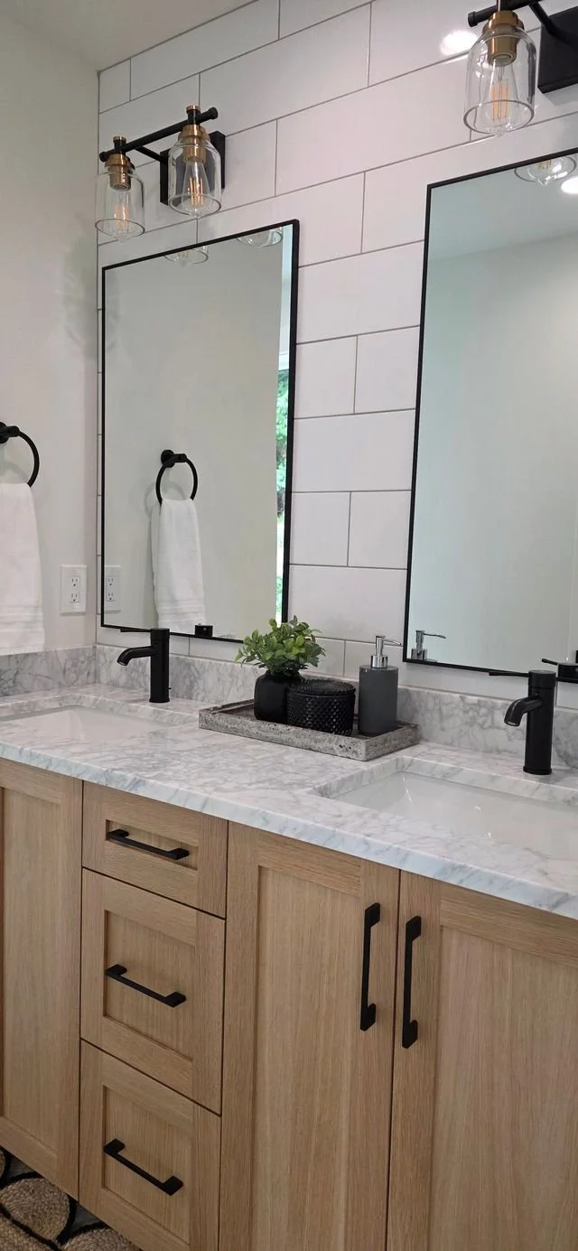A modern bathroom vanity with a white marble countertop, twin mirrors, black fixtures, and a decorative tray with a plant and soap dispenser, lit by industrial-style light fixtures.