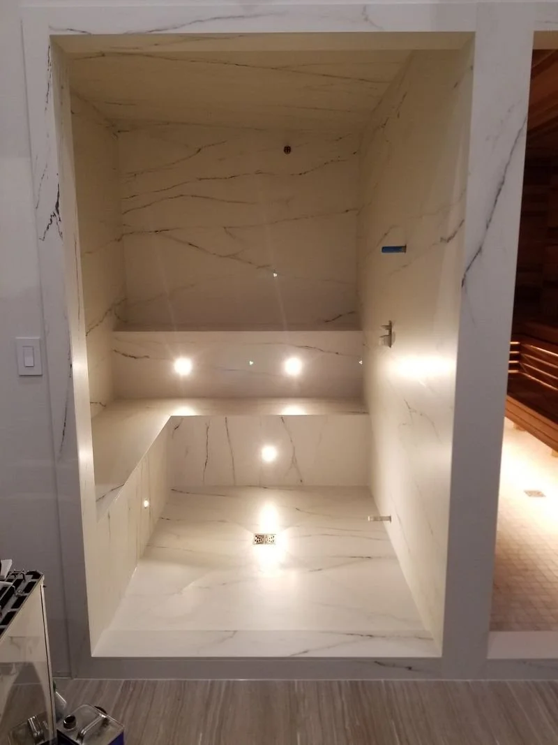 Marble-tiled shower with overhead lighting and a drain at the bottom.