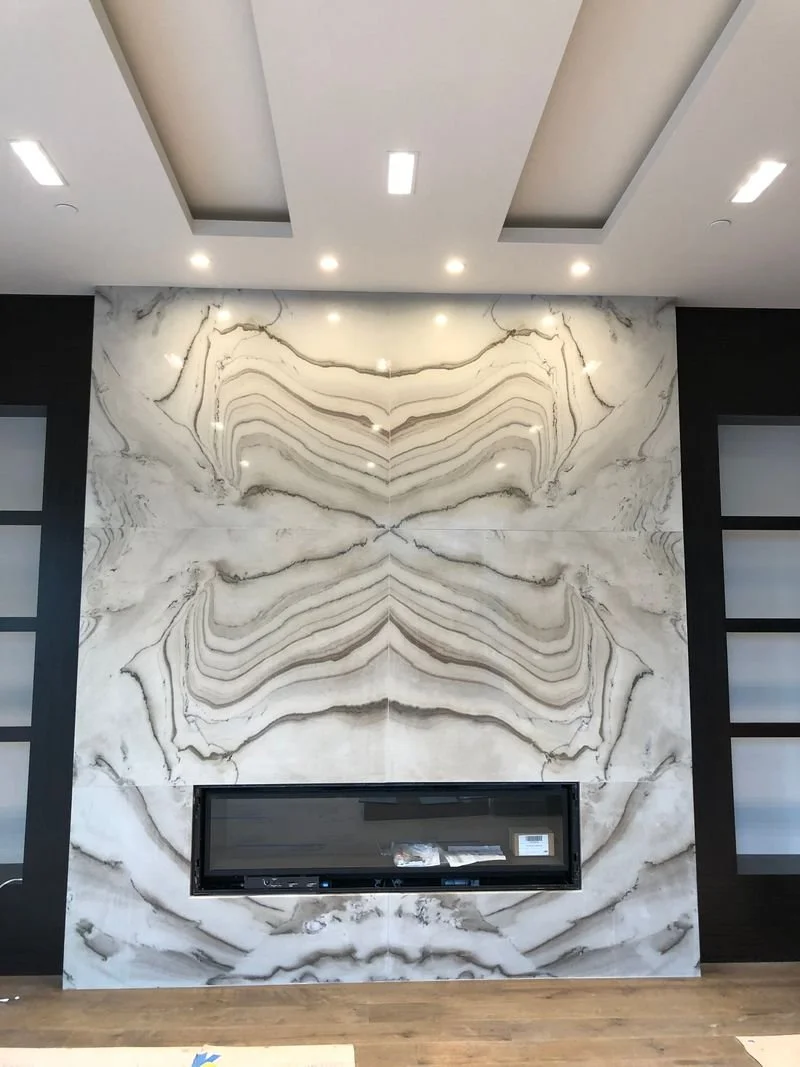 Interior of a modern room with a large marble fireplace with symmetrical natural patterns, flanked by dark wooden panels with glass shelving, and a wooden floor.