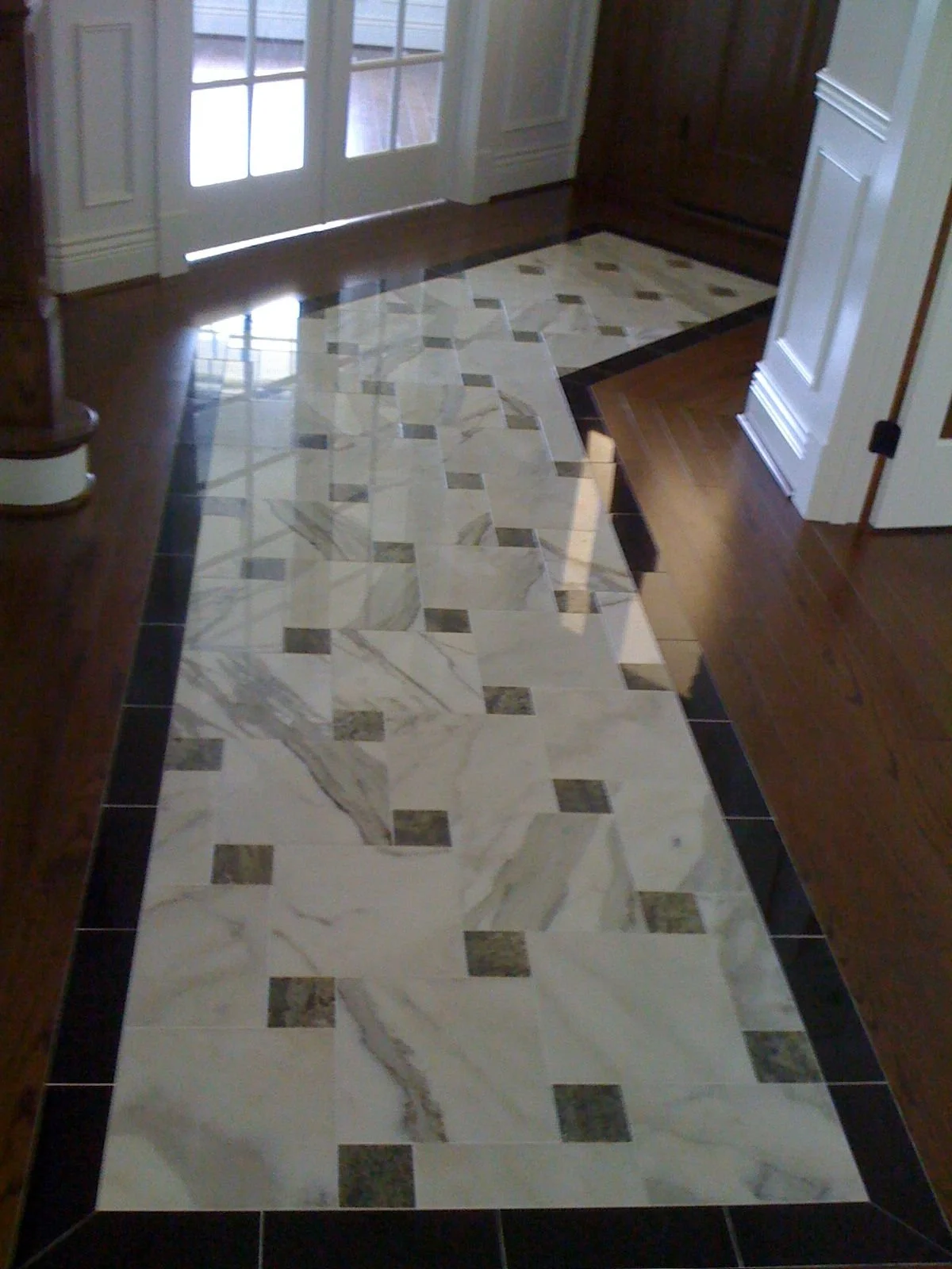 Interior view of a room with a decorative marble tile floor design and wooden flooring around it, glass double doors at the background.