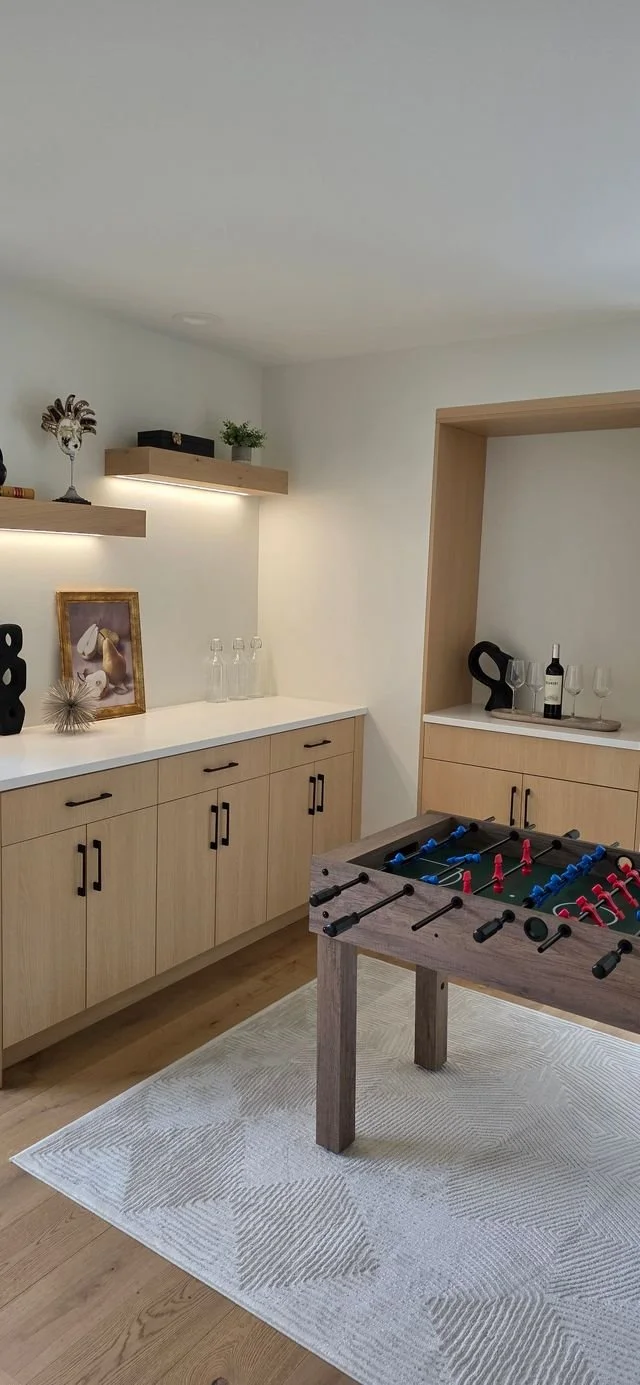 A room with a foosball table on a white rug, wooden cabinets, decorative shelves, and various items including glasses, wine bottles, artwork, and decorative objects.
