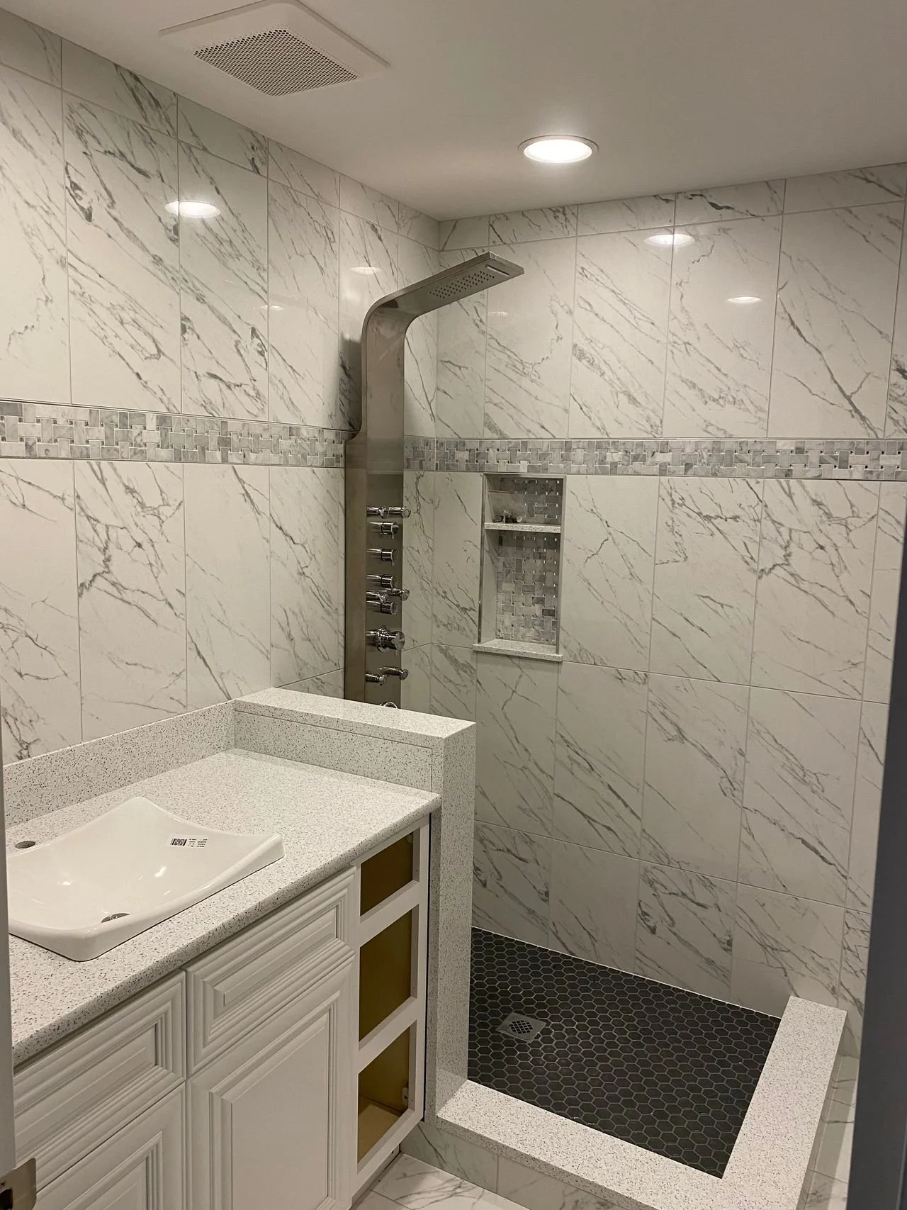 Bathroom with marble tiles, a small walk-in shower with black hexagonal tiles on the floor, a built-in shelf, and a white vanity with a vessel sink.