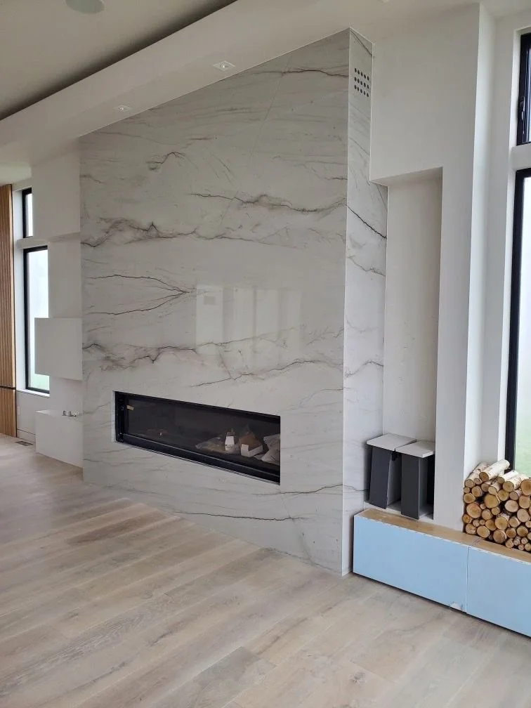 Modern living room with a marble fireplace wall, light wood flooring, split windows, and a bundle of logs stored next to a blue storage box.
