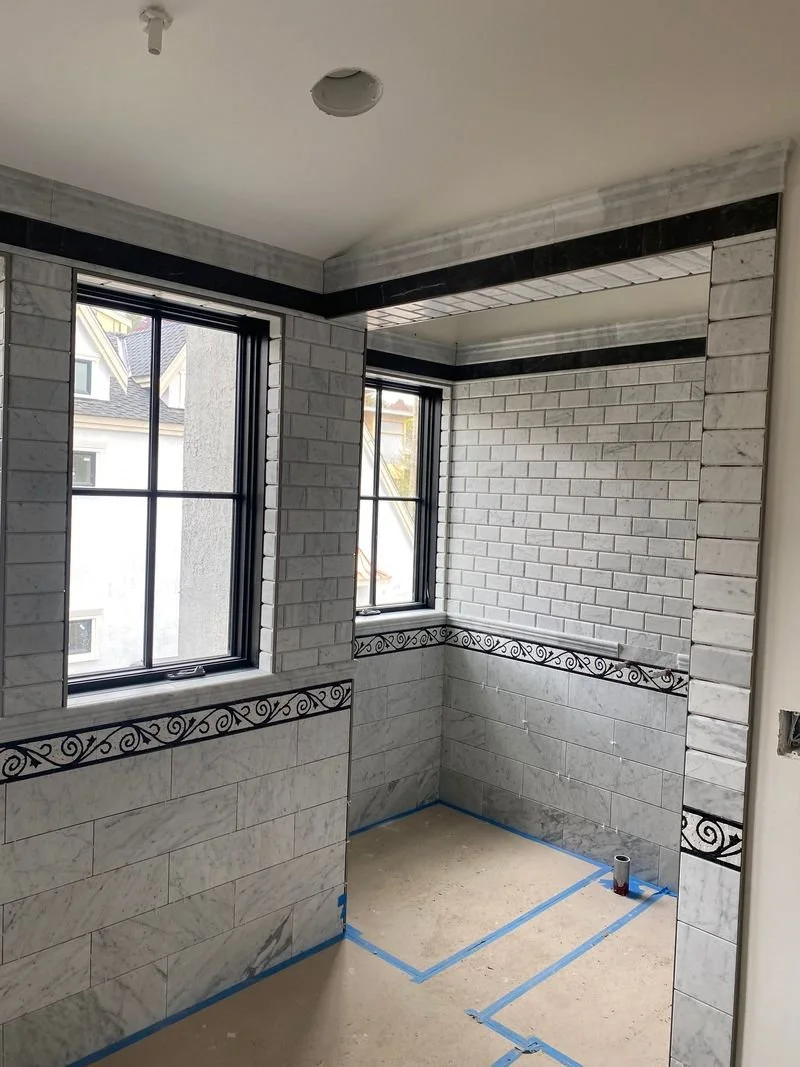 Interior of a room under construction with white brick walls, black decorative tile accents, two black-framed windows, and a concrete floor marked with blue painter's tape.