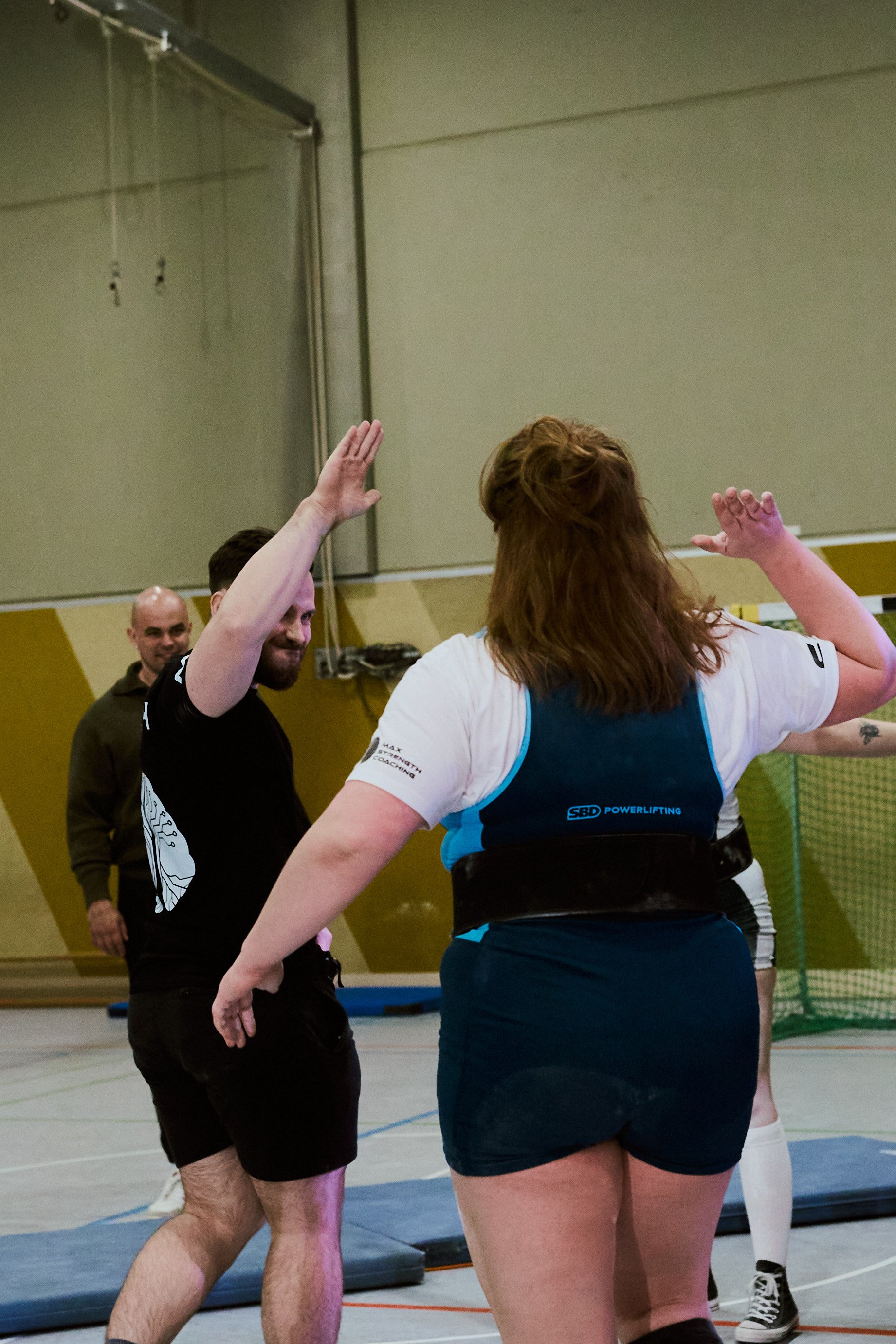 Zwei Personen beim Powerlifting in einem Turnsaal, die sich zum High Five begrüßen, eine ist Mann, die andere Frau, im Hintergrund eine dritte Person.