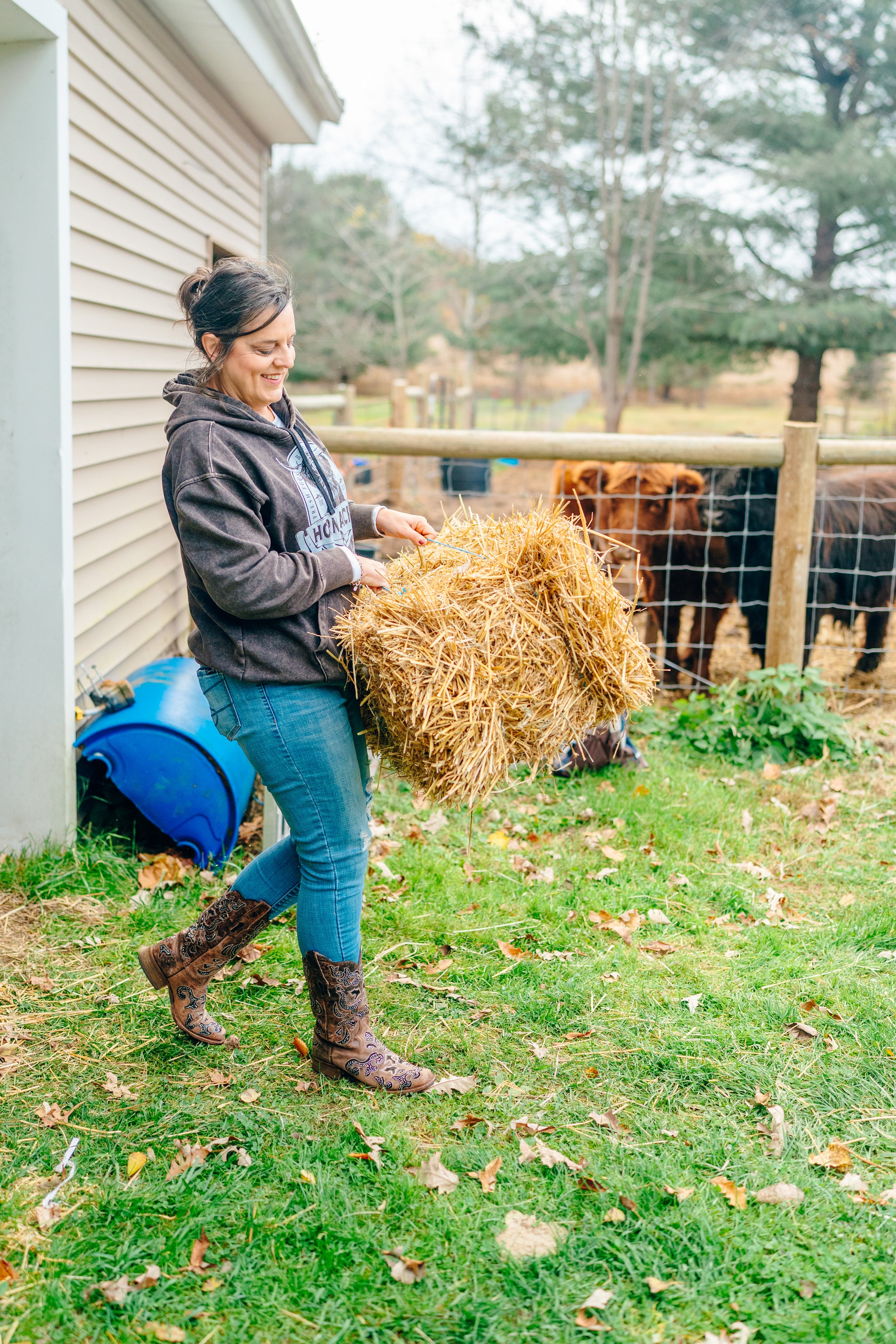 A woman in cowboy boots and a hoodie is holding a bundle of hay outside near a fence with cows.