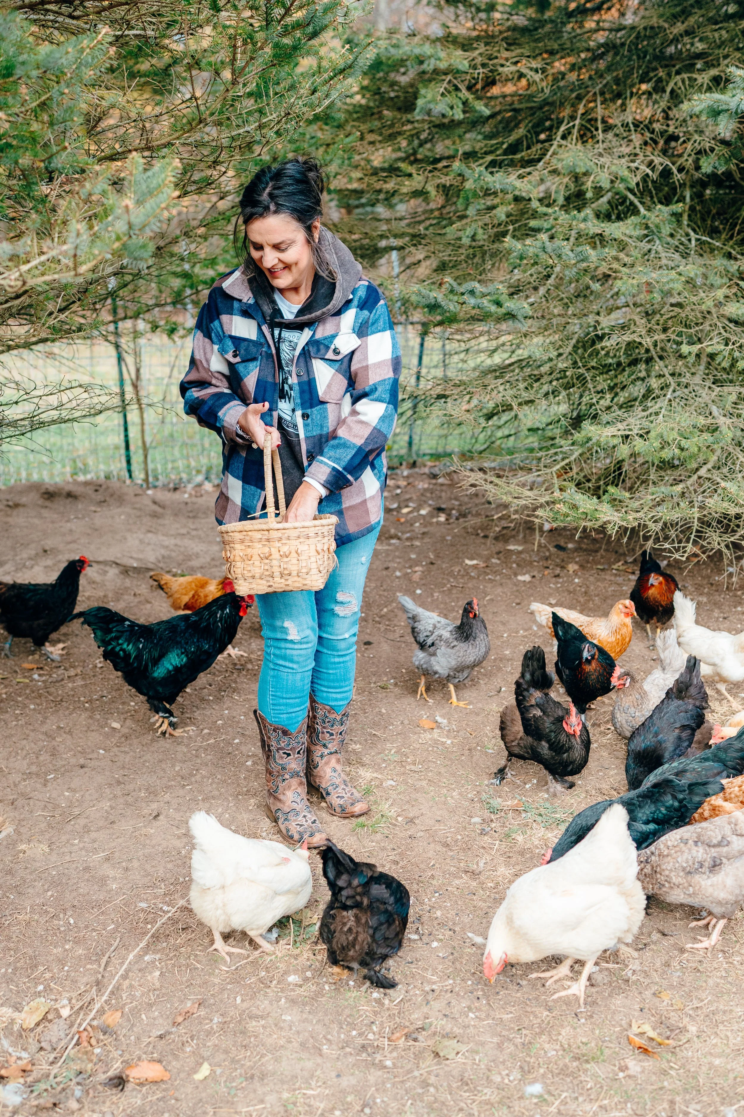 A woman in plaid jacket and boots feeds chickens in a backyard with trees.