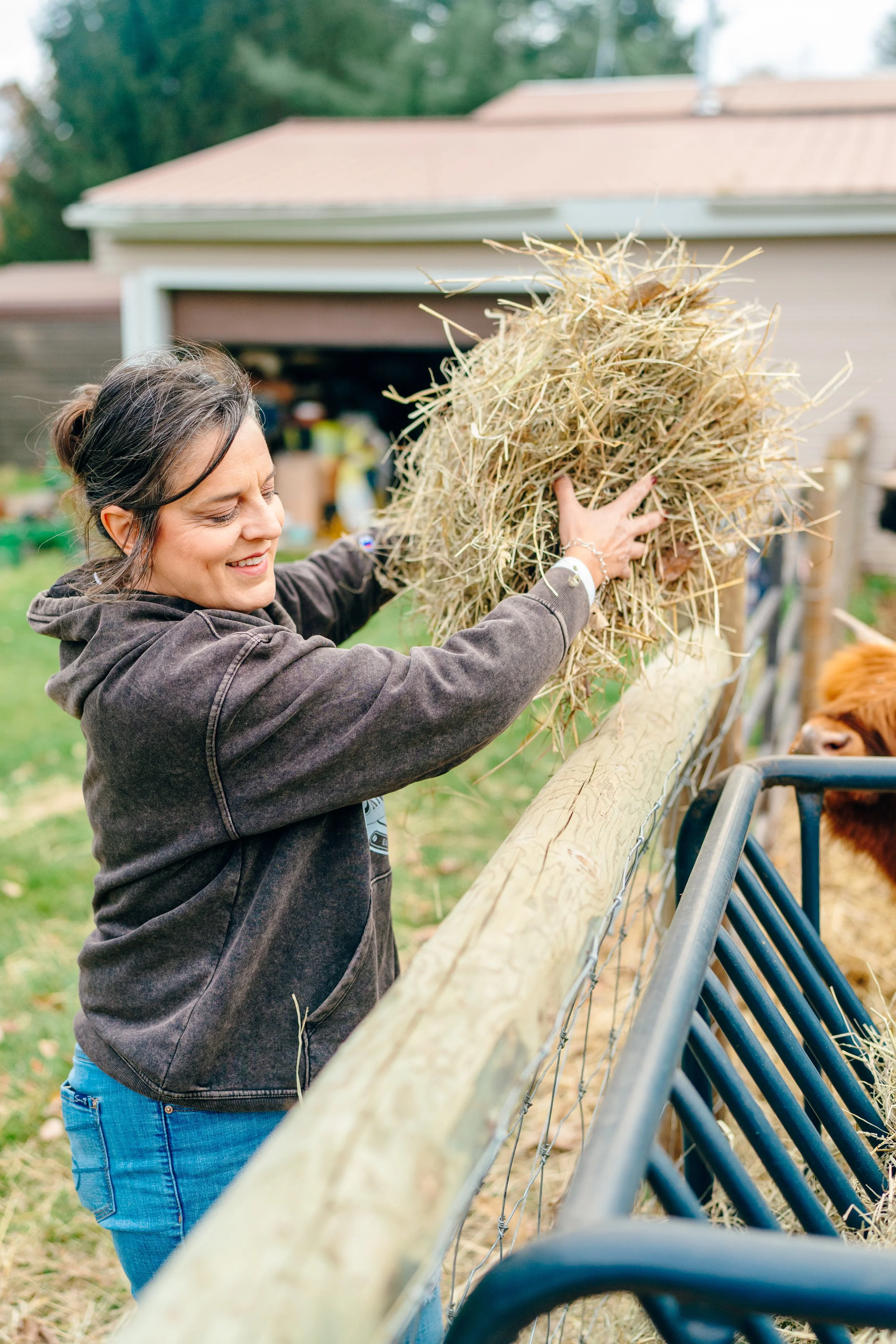 A woman smiling and holding a large bundle of hay while standing next to a fence, with a cow behind the fence and a garage with various items in the background.