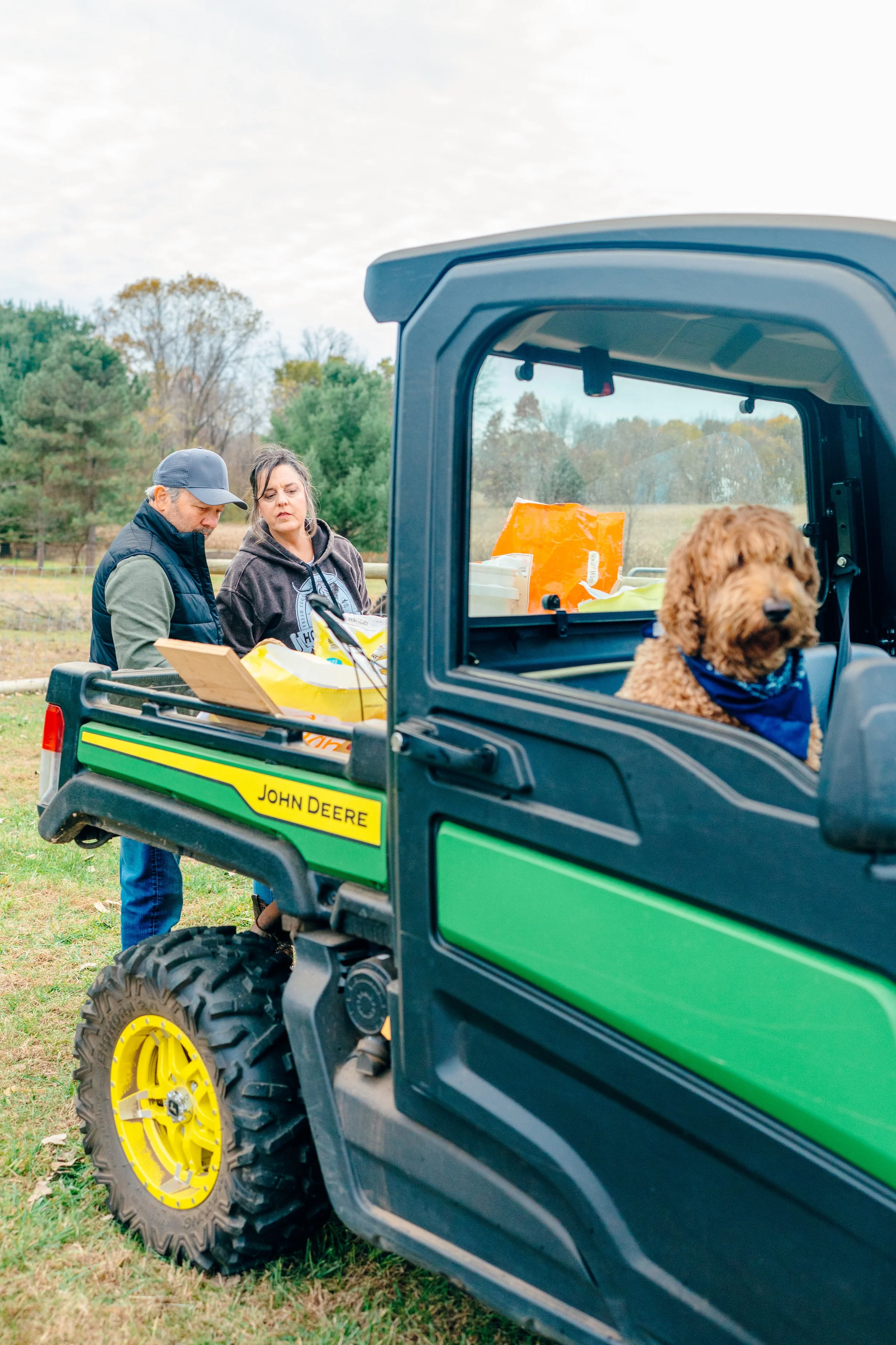 Two people and a dog near a green John Deere utility vehicle in an outdoor setting, with trees in the background.