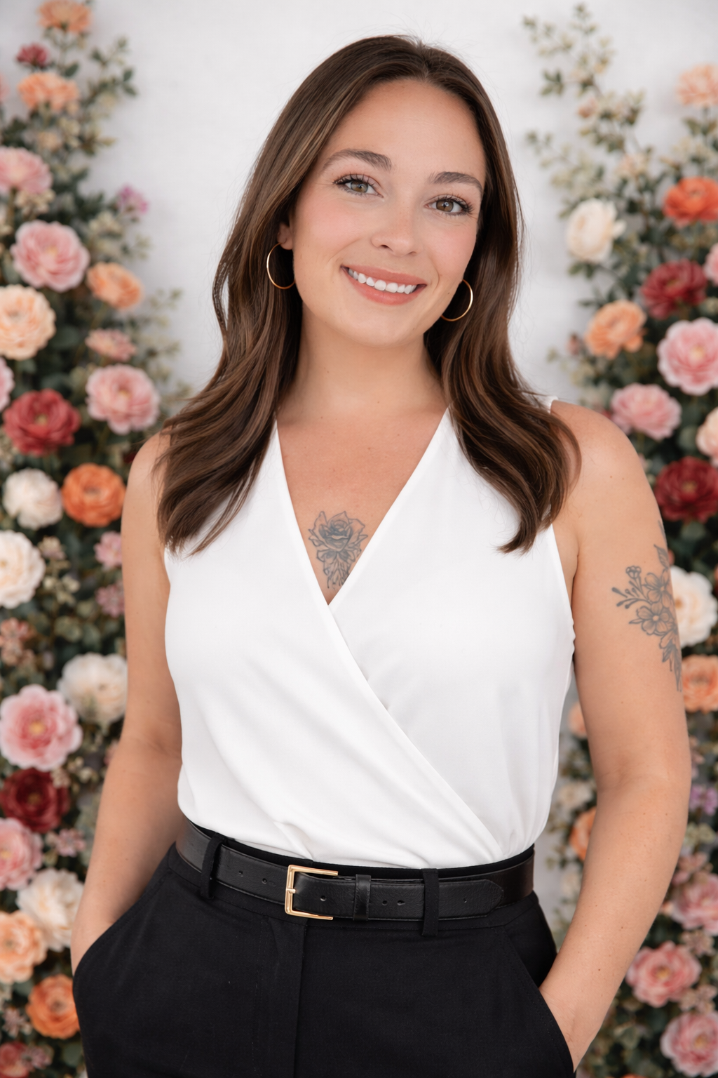 A young woman with shoulder-length brown hair, gold hoop earrings, and tattoos on her chest and arm, smiling in front of a backdrop of pink, white, and orange flowers.