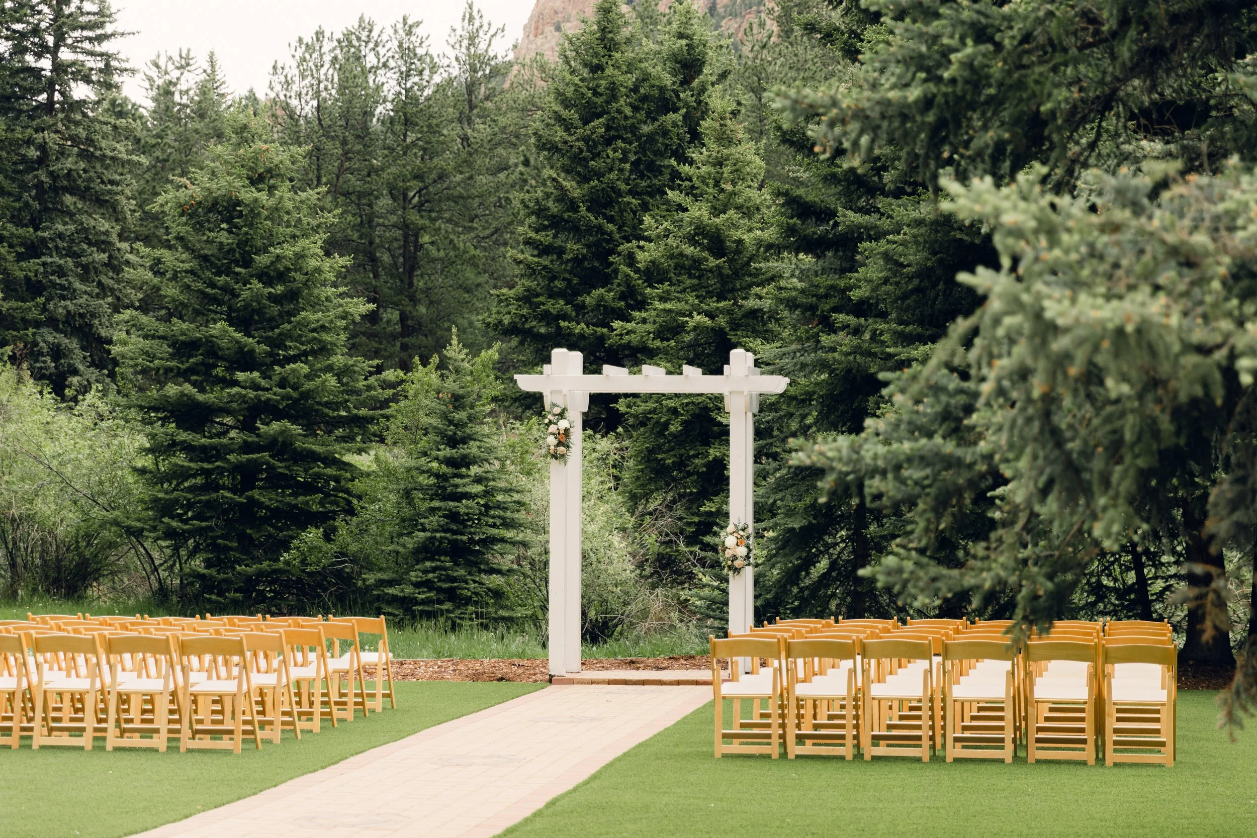 Outdoor wedding ceremony setup with a white arch decorated with flowers, wooden chairs arranged in rows on a grassy area, and a background of tall green trees.